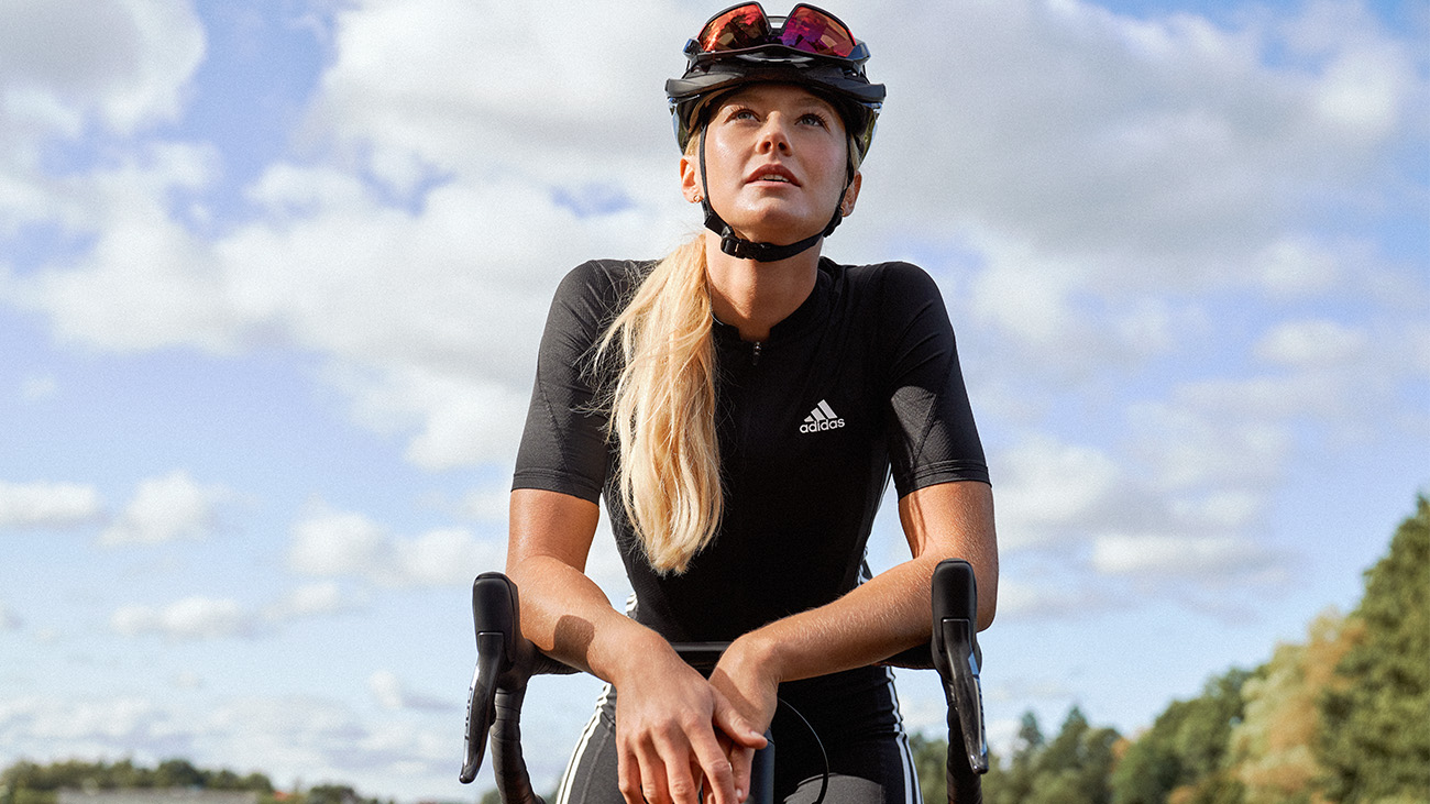 Eine Frau mit Helm in Radsportkleidung lehnt sich über den Lenker und macht am Straßenrand Pause.