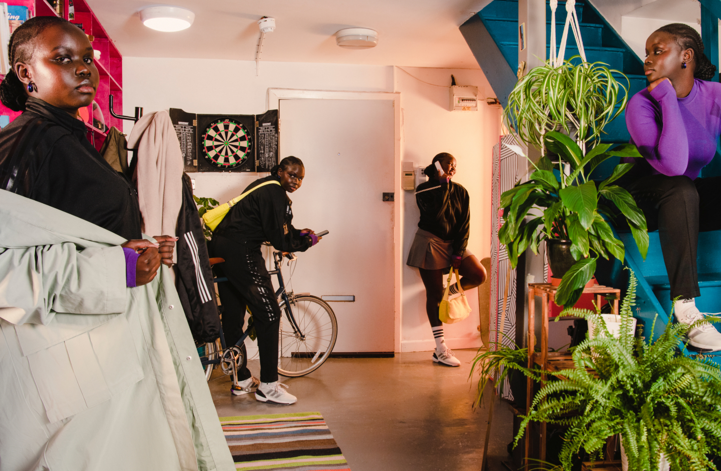 Young woman in four poses across living room backdrop: facing camera modelling long coat, holding bike in hand wearing adidas track suit, talking on a landline phone with foot pressed against wall in adidas track jacket, and sitting on stairs looking off camera in adidas sportswear top and pants.