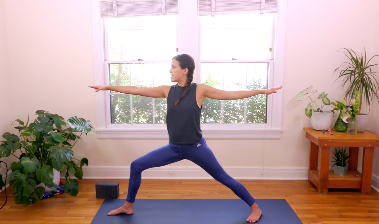 Framed by plants, Adriene is seen in her studio holding the warrior II pose.