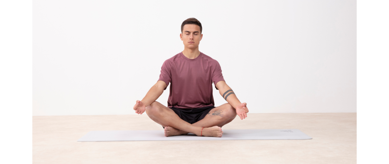 Un hombre en una postura de yoga medita con las piernas cruzadas y las manos apoyadas sobre las rodillas. Lleva una camiseta morada y un pantalón corto negro, todo de adidas.