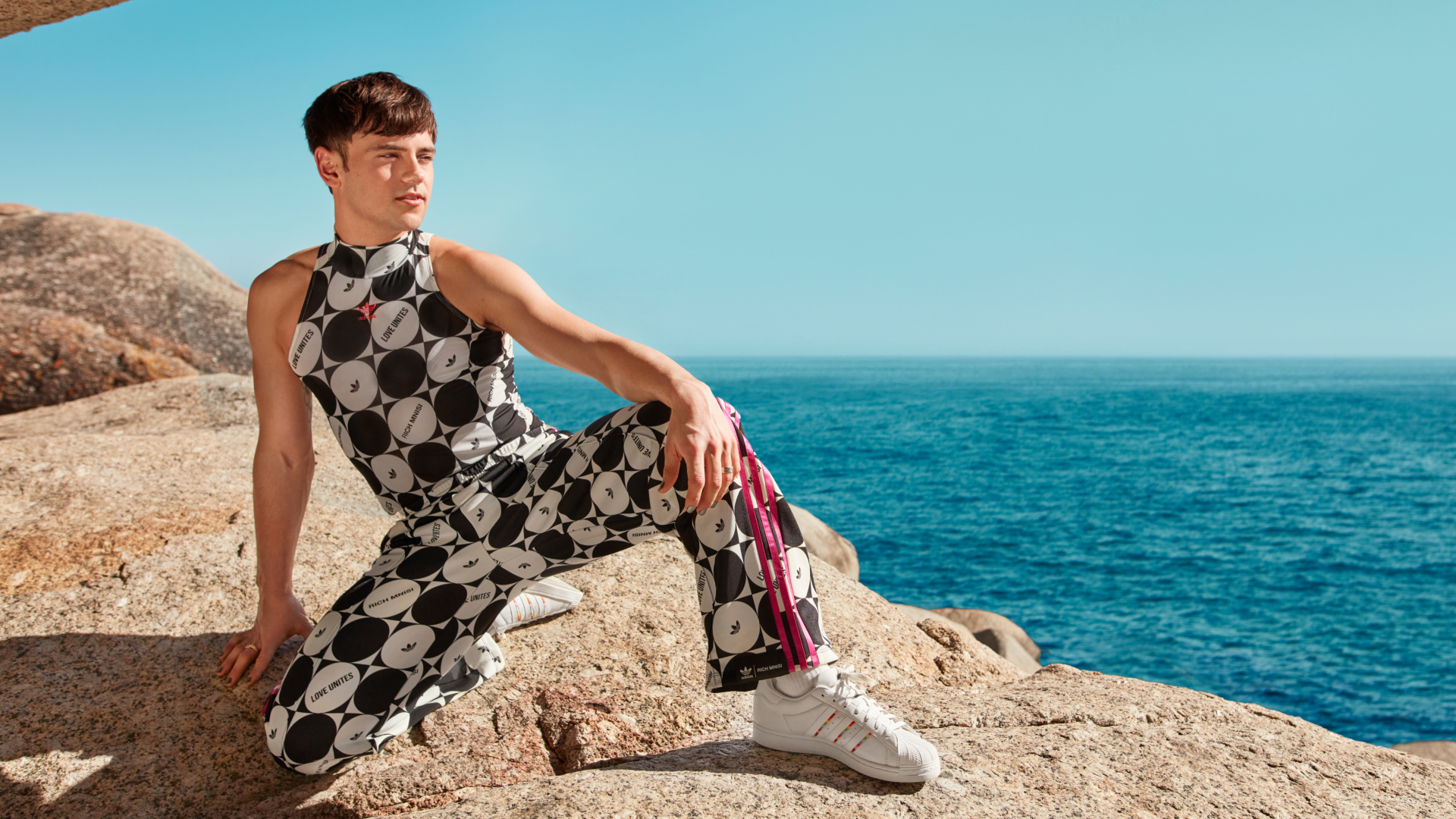 Tom Daley posa en una gran roca con un cielo y un océano azules e infinitos de fondo, y luce prendas de la colección Pride de adidas x Rich Mnisi.