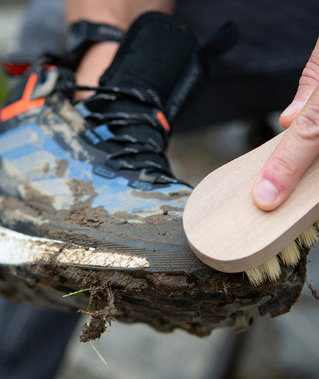 Primer plano de una bota de montaña adidas TERREX con la parte superior embarrada y la mano de alguien limpiándola con un cepillo.