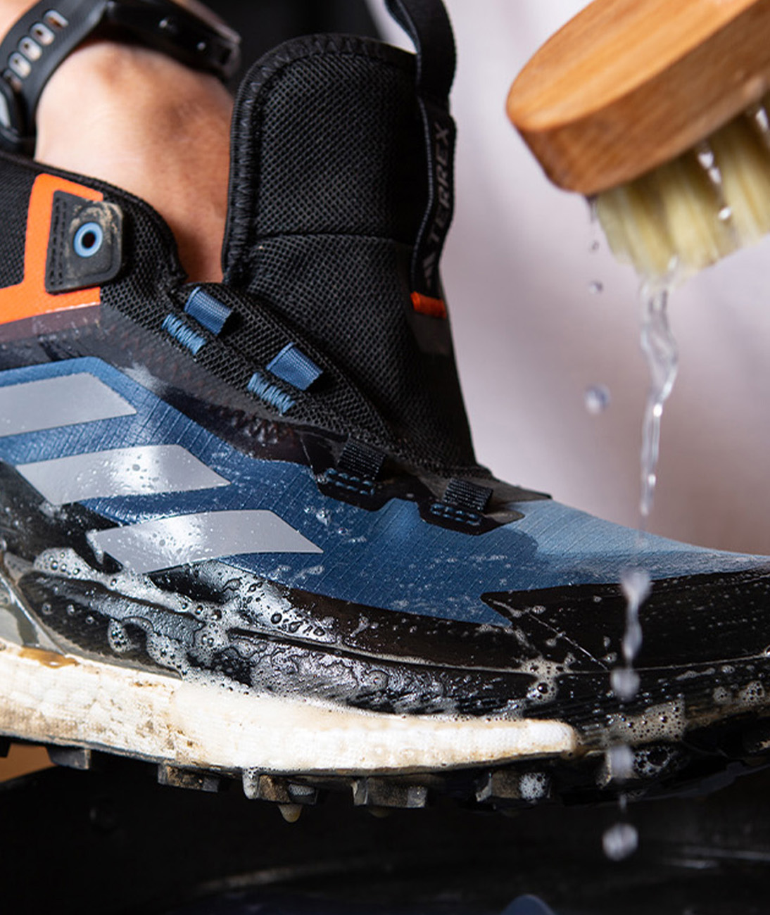 Una bota adidas TERREX mojada sobre un recipiente con agua jabonosa y la mano de alguien que la frota con un cepillo.