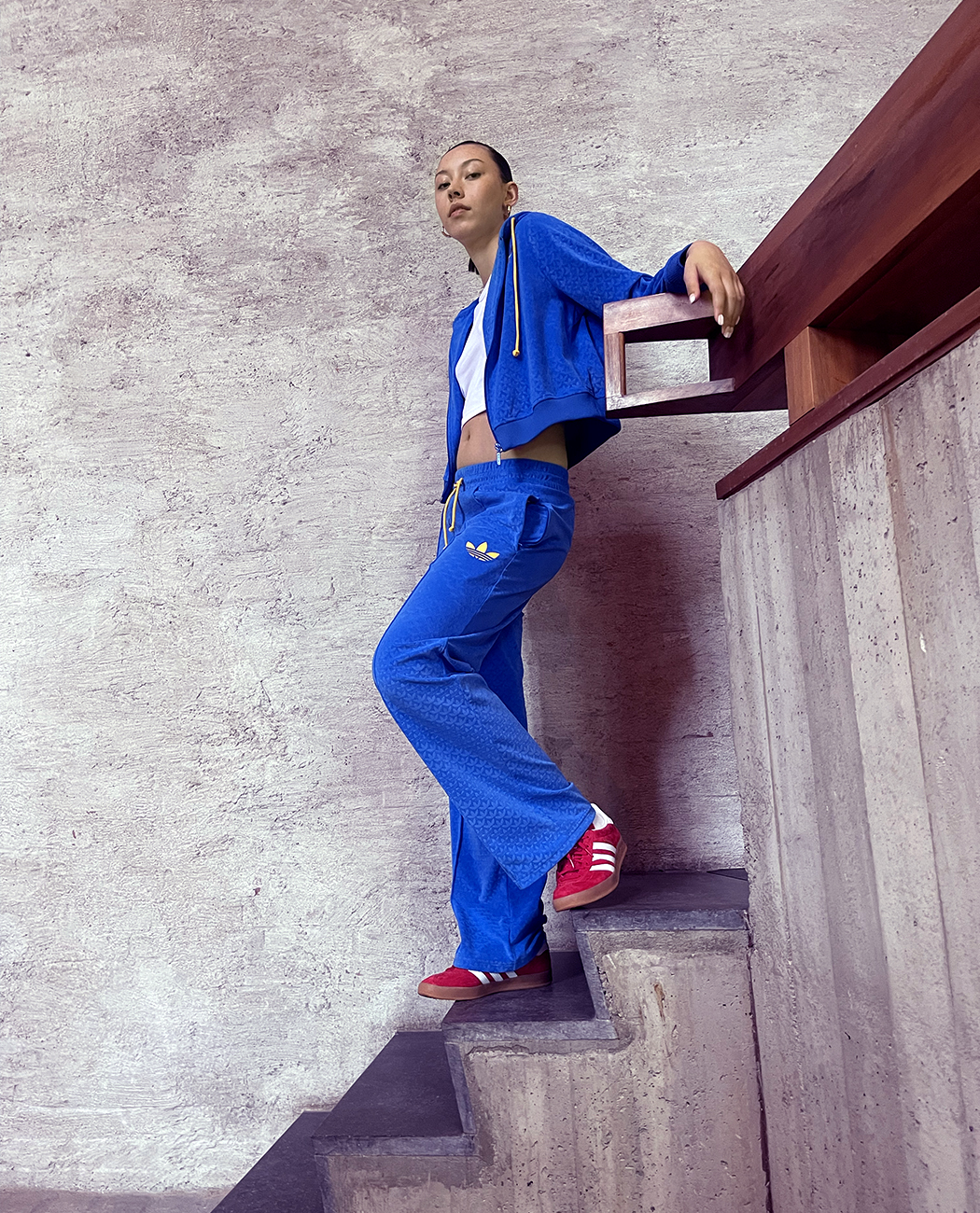 Female model standing on a flight of stairs wearing a blue outfit on a white t-shirt and red adidas Gazelle sneakers with with stripes.