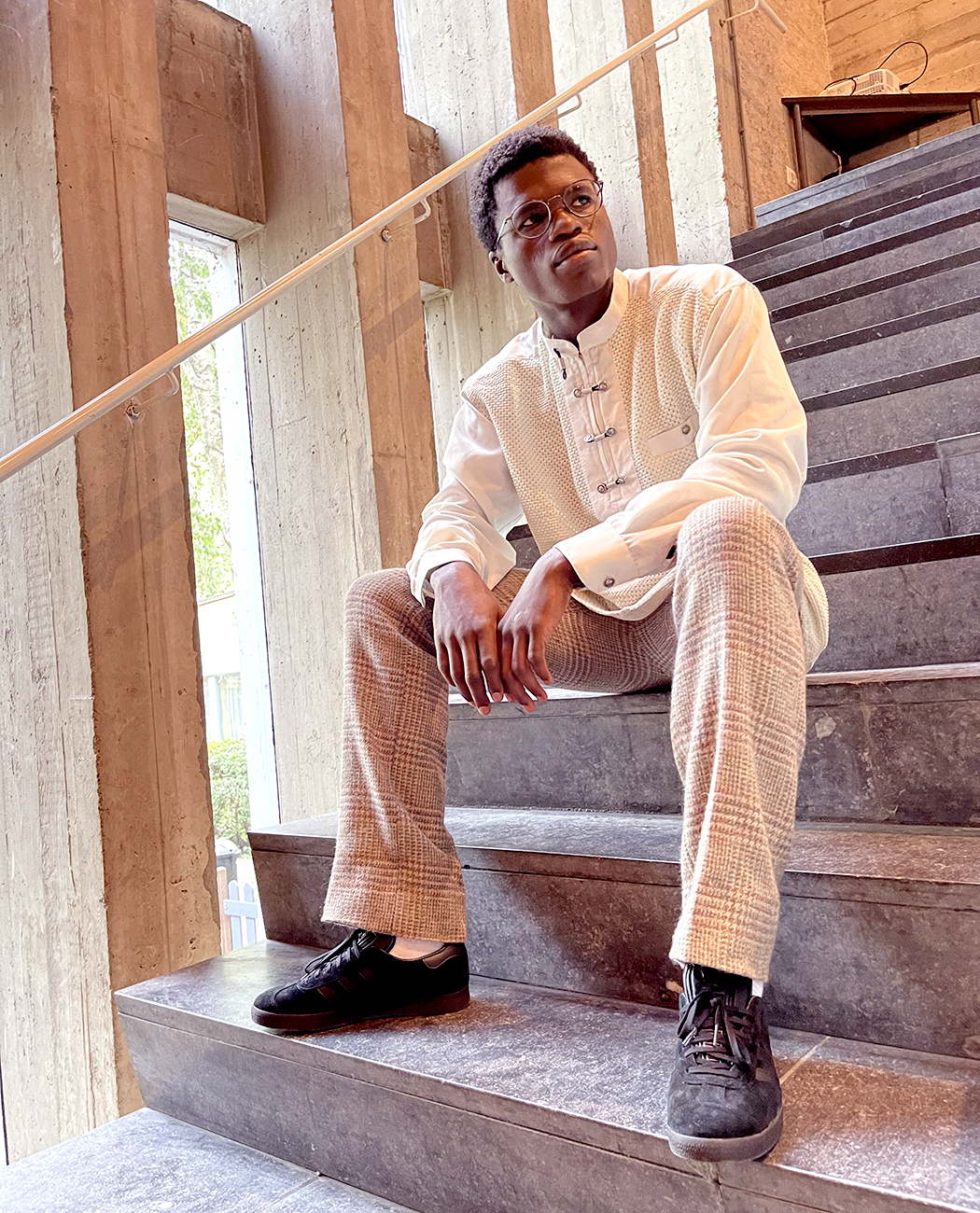 A male model sitting on a flight of concrete stairs whilst wearing an off-white and beige outfit and all black adidas Gazelle sneakers.