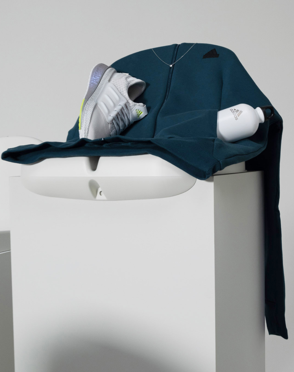 Zdjęcie bluzy z kapturem zapinanej na zamek, butelki na wodę oraz butów treningowych X_PLRBOOST z kolekcji ubrań sportowych adidas Z.N.E. w aranżacji na plastikowym siedzeniu na trybunach stadionu.