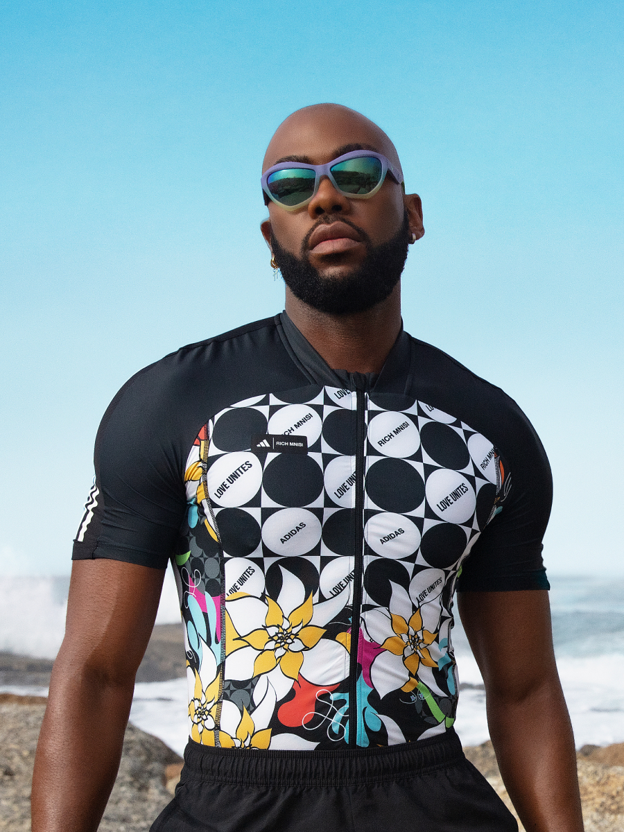 Rich Mnisi em pose contra um oceano azul-claro como imagem de fundo, com uma blusa preta e branca da coleção adidas x Rich Mnisi Pride Collection.