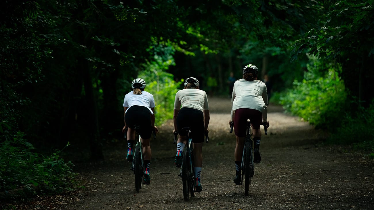 Three cyclists ride side by side on a gravel path in a forest