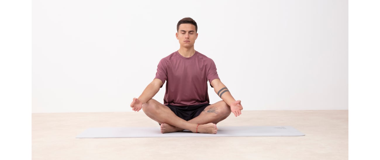 A man meditates in a yoga pose with crossed legs and hands resting on his knees, wearing a purple adidas t-shirt and black shorts.