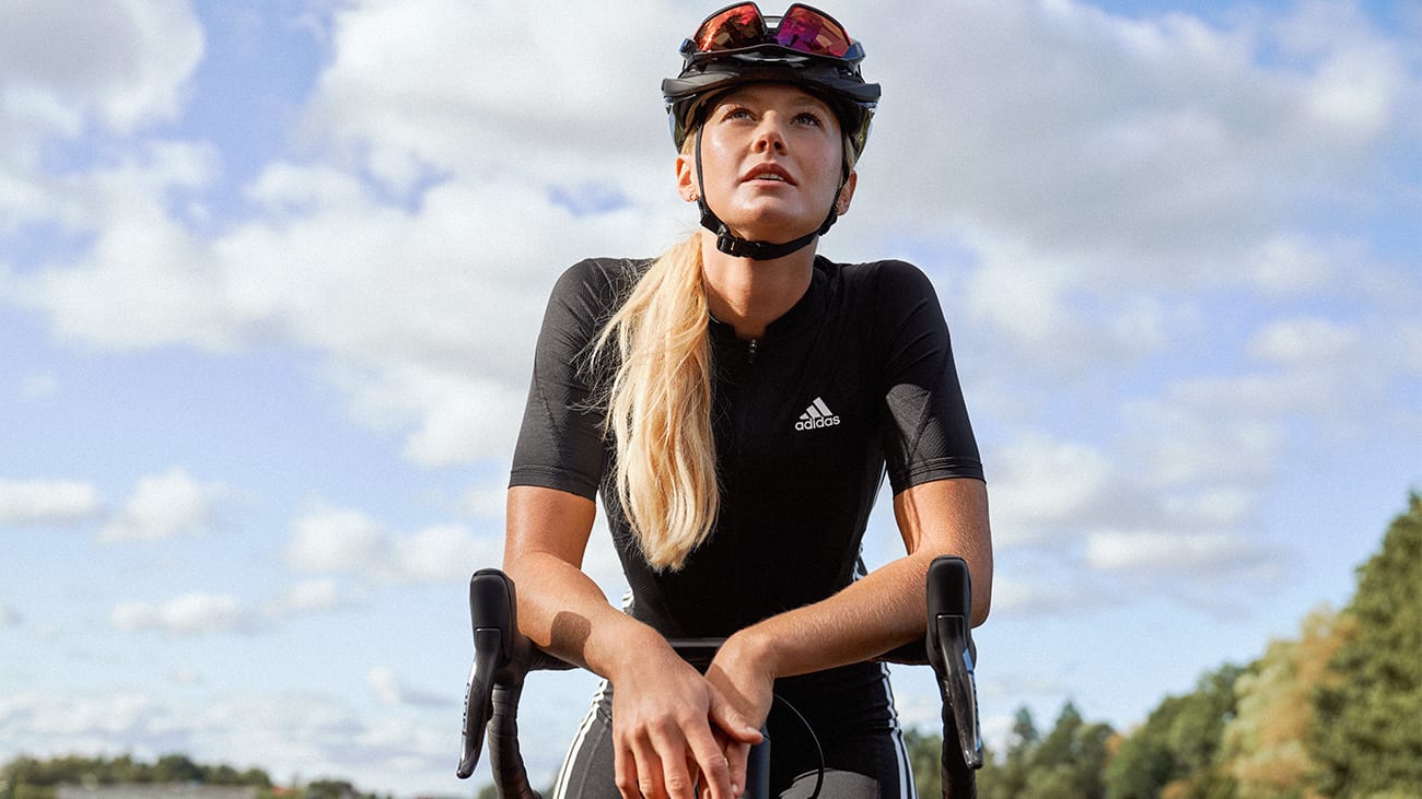 fw21-cycling-road-body-2 A woman wearing cycling clothing and a helmet leaning on her bike resting in the road