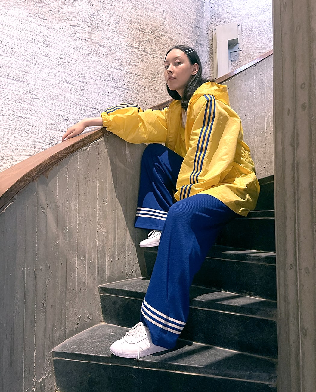 Female model sitting on a flight of stairs wearing blue pants and a yellow jacket, with white adidas Gazelle sneakers.