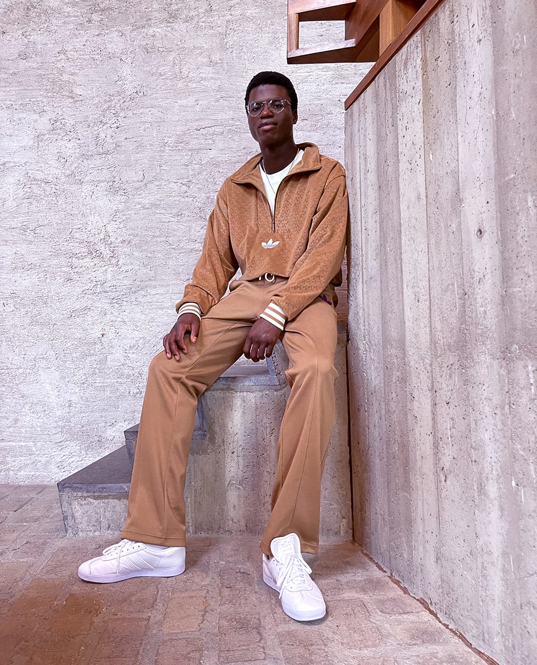 A male model sitting on a concrete block whilst wearing a brown adidas sweater and brown pants with all white adidas Gazelle sneakers.