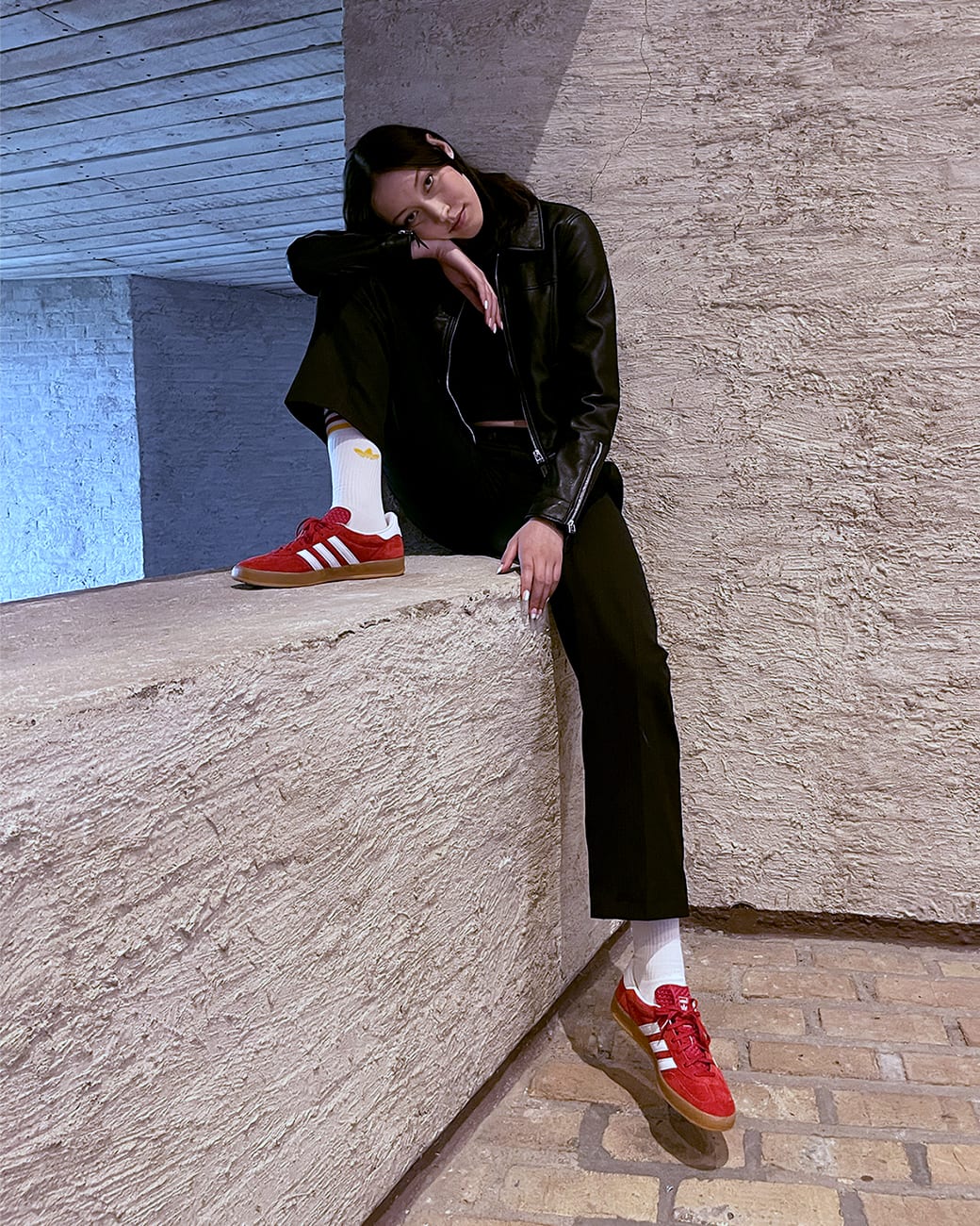 A female model sitting on a concrete block whilst wearing an all black outfit, white socks and red adidas Gazelle sneakers with white stripes.