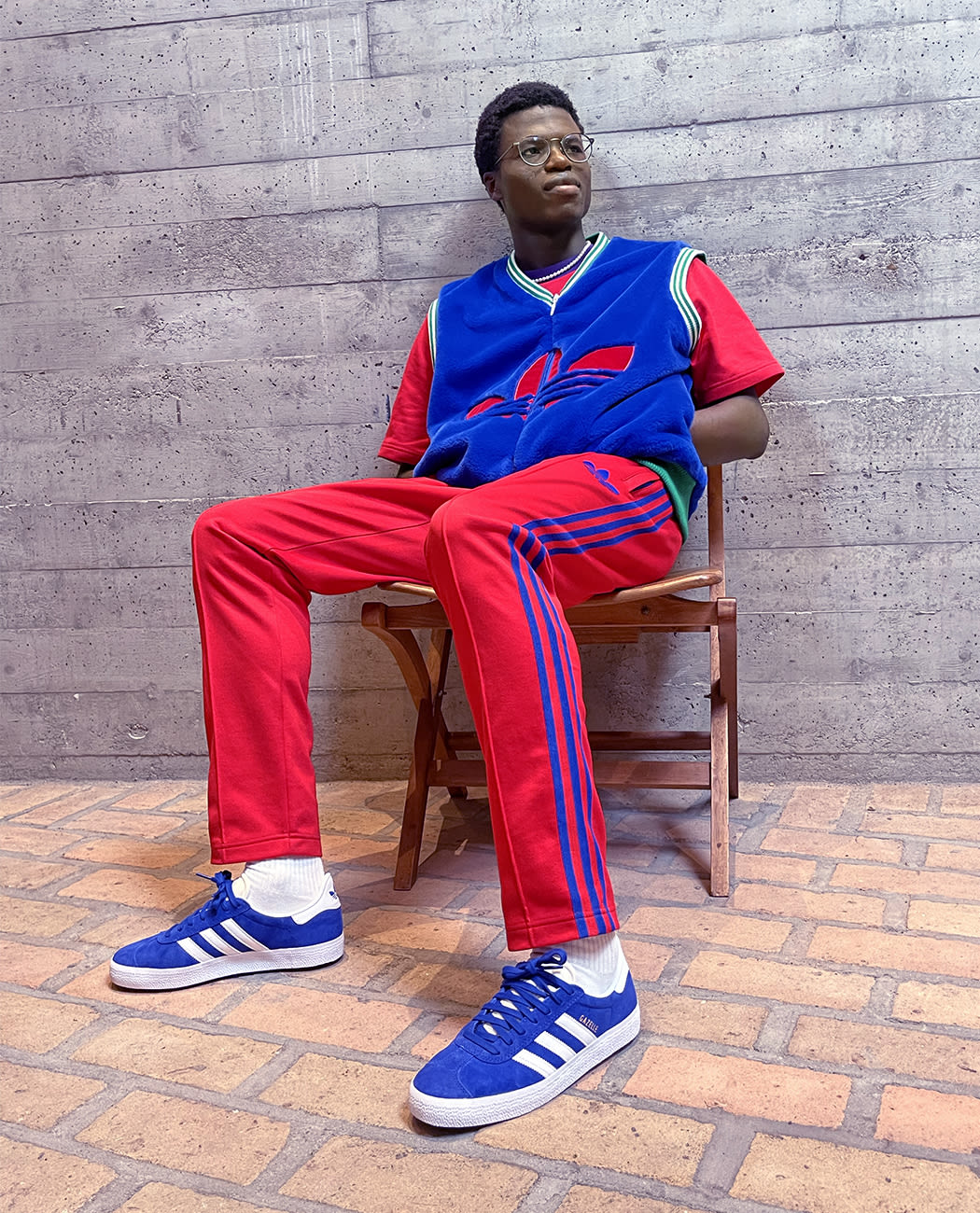 A male model sitting on a chair against a concrete wall, wearing red pants, a blue adidas vest and lightblue adidas Gazelle sneakers with white stripes.