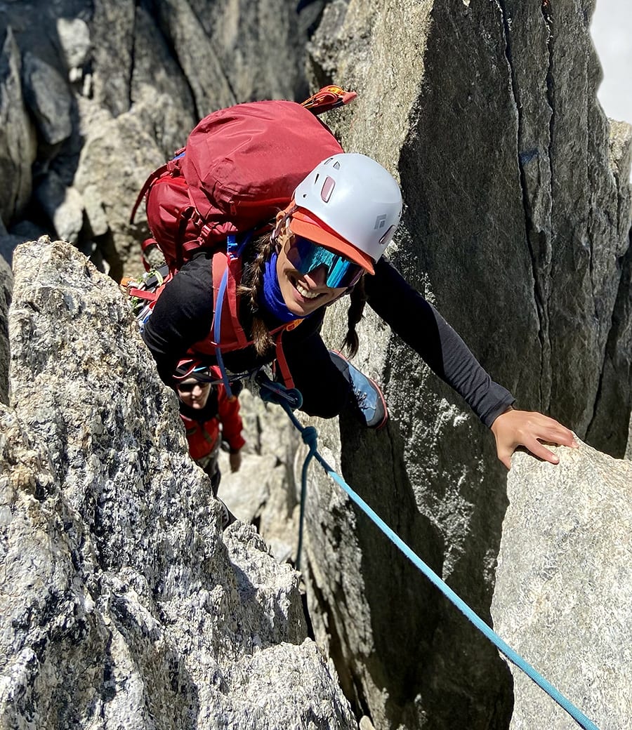 Female mountaineer wearing a white helmet smiles as she approaches the top of a rocky crag