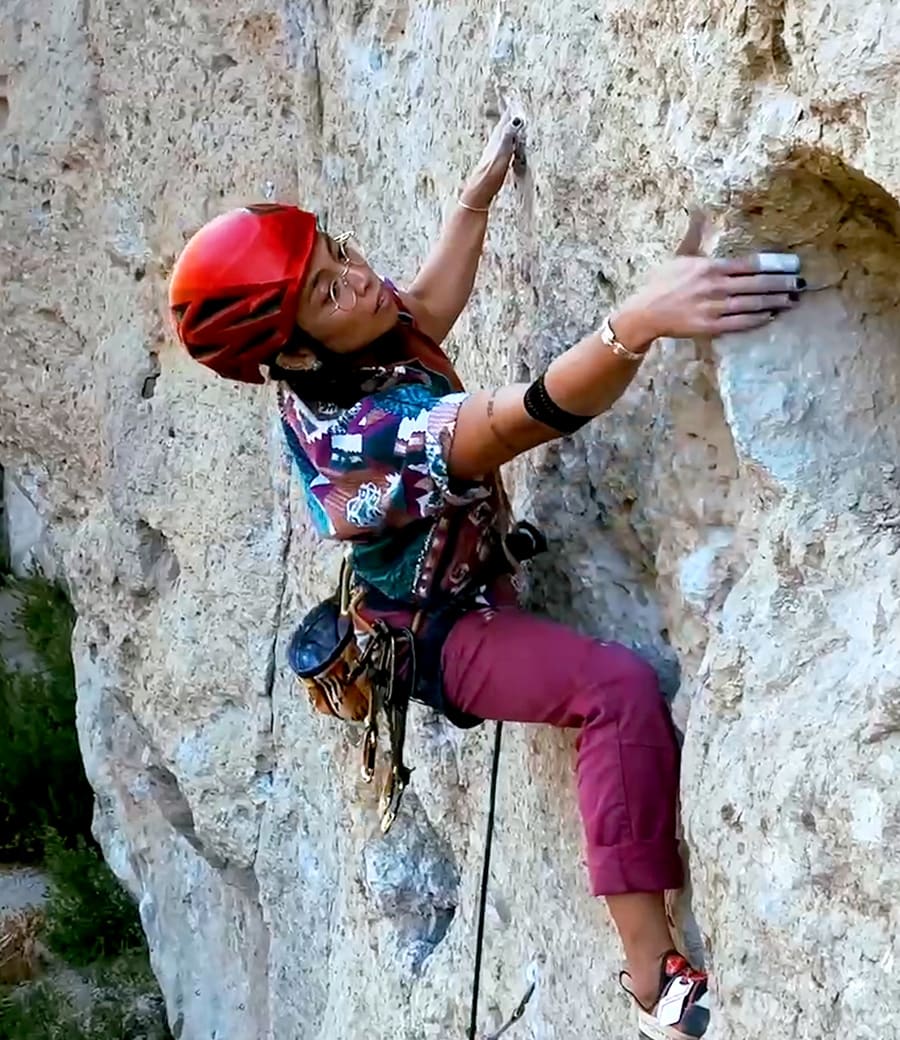 Female climber on a rock face wearing a red helmet and bright shirt reaches out for a hand hold