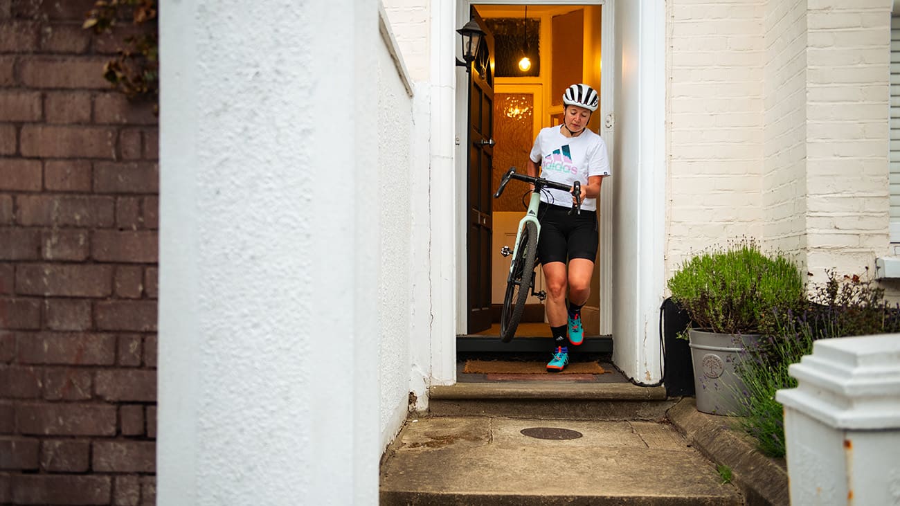 A woman dressed in cycling gear carries her bike out of her house through the front door