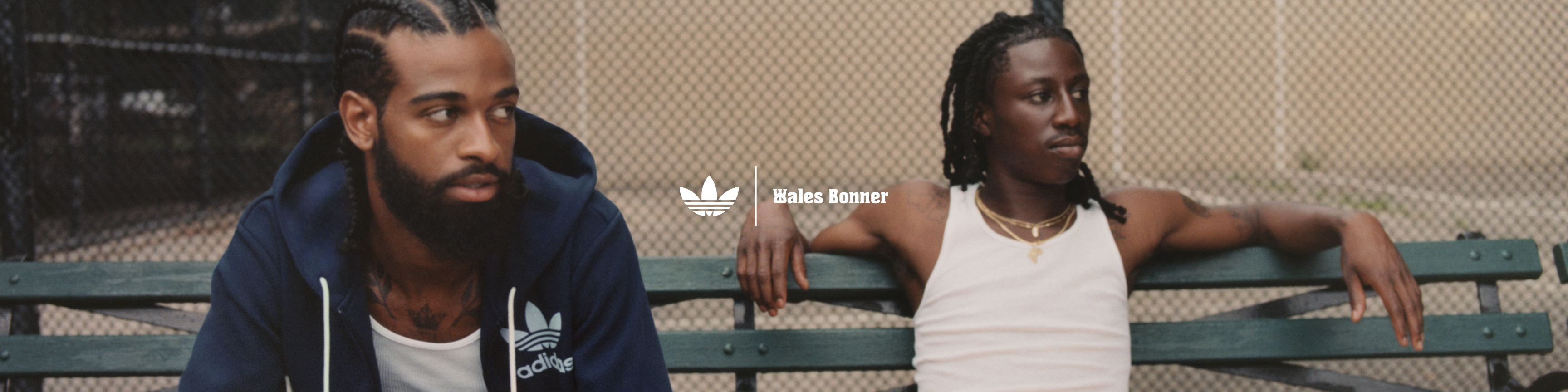 Two models pose on a park bench, with a handball court in the background, wearing the adidas Originals by Wales Bonner collection.