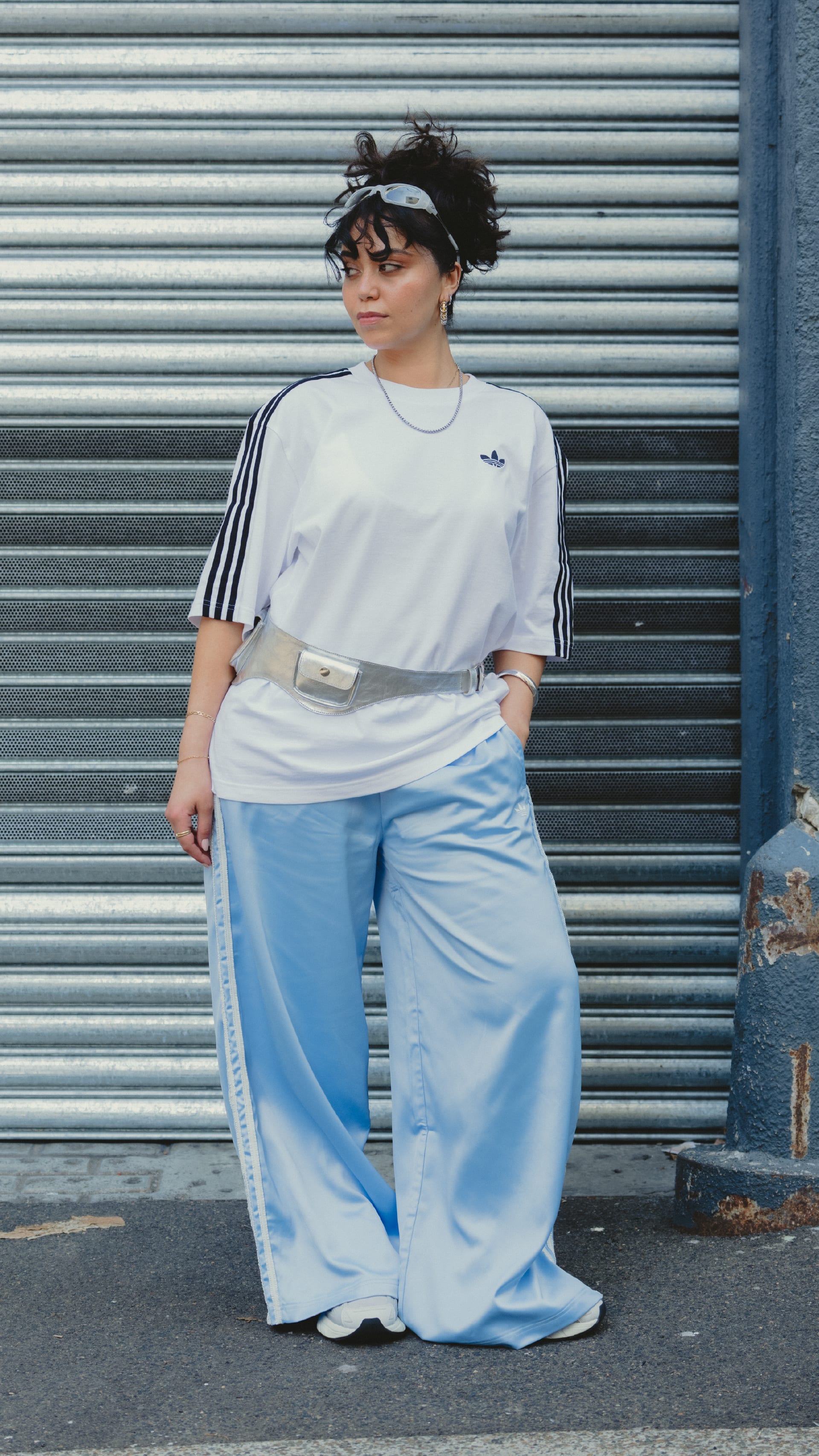 A person wearing a white t-shirt and blue wide-leg pants stands in front of a metal shutter backdrop.