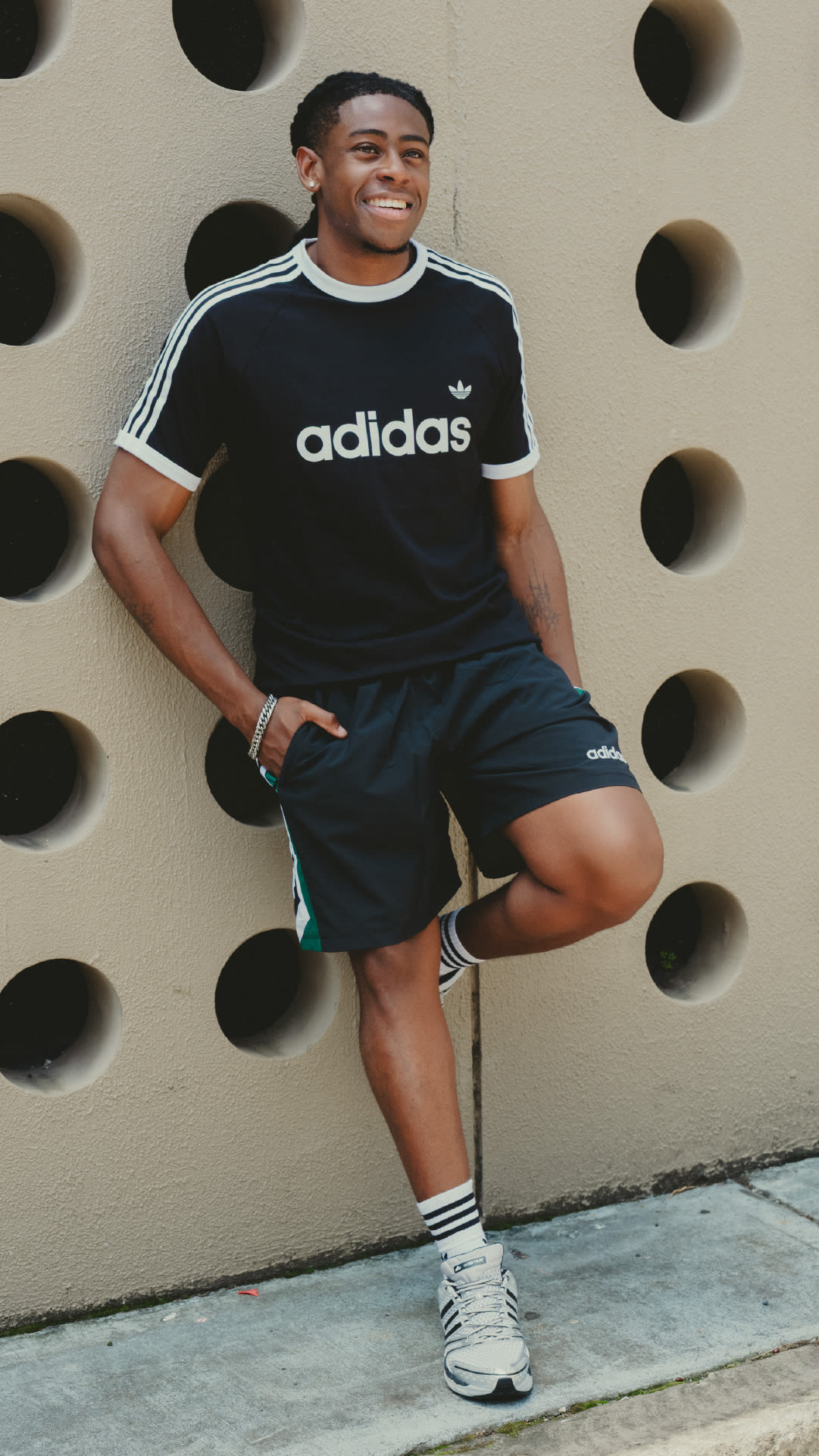 A smiling young man wearing an Adidas t-shirt and shorts, standing in front of a wall with circular patterns.