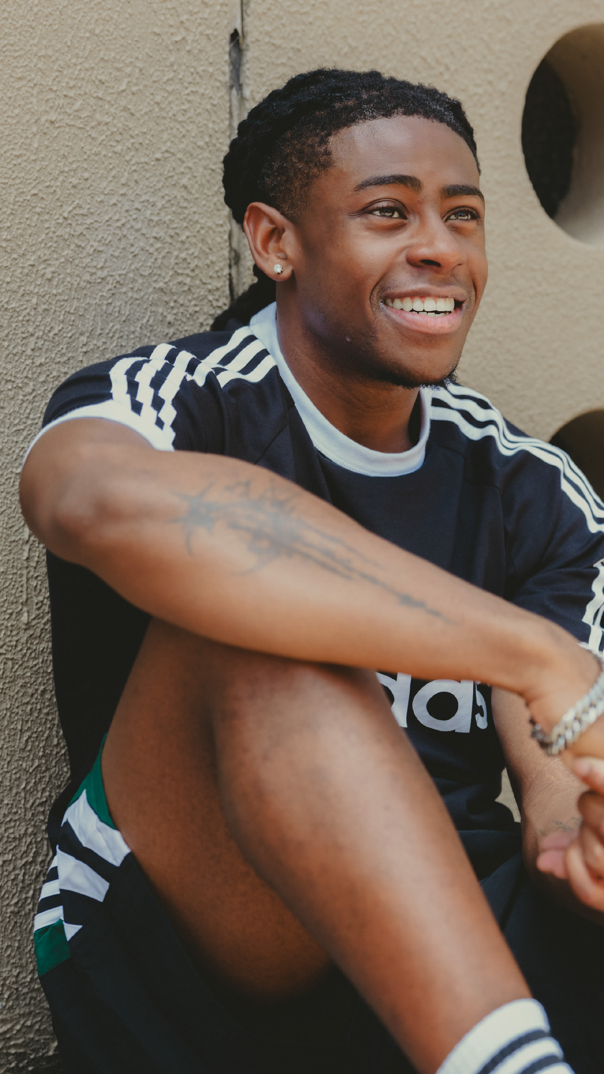 A young man with a warm smile, wearing a black and white striped shirt, sits against a plain wall background.
