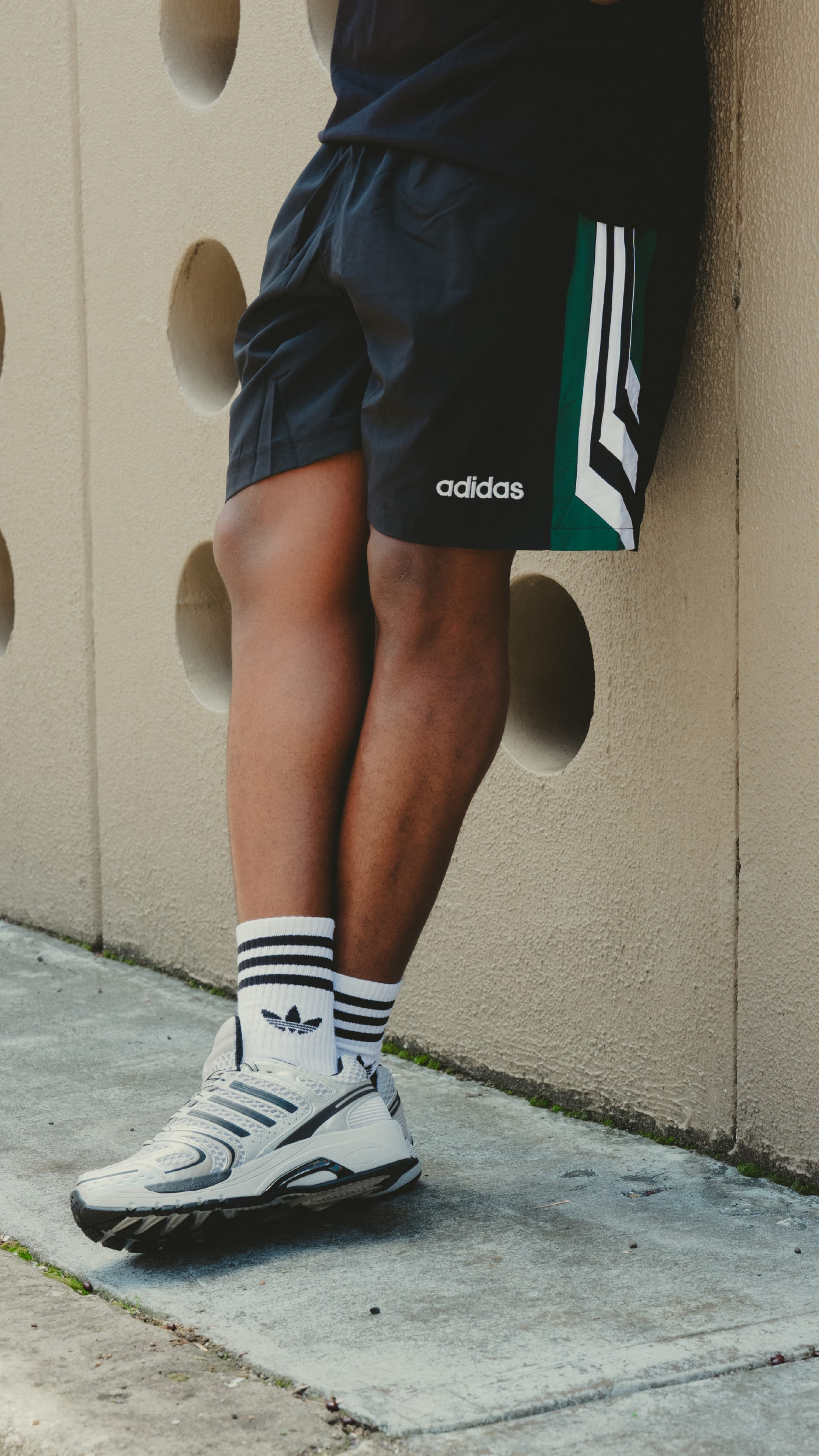 The image shows a person's lower body wearing Adidas athletic shorts and shoes, standing in front of a concrete wall with circular openings.