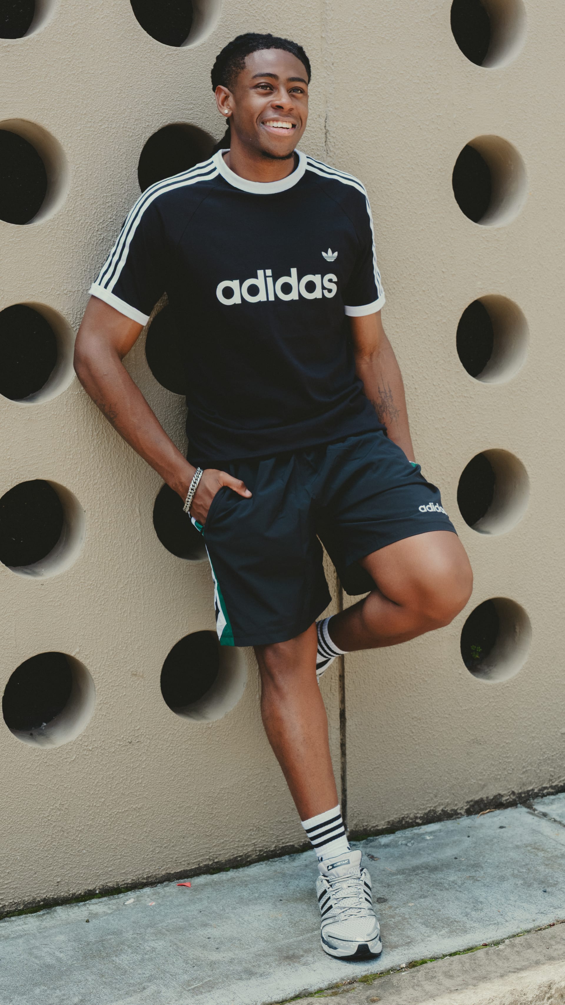 A smiling young man wearing an Adidas t-shirt and shorts stands in front of a wall with circular patterns.