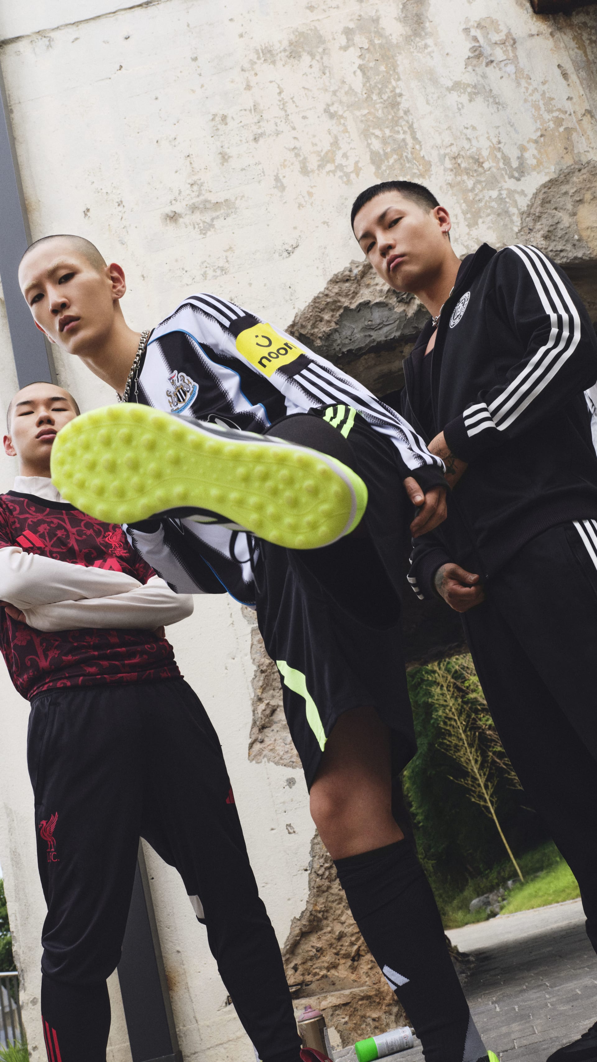 The image shows two young men wearing black athletic clothing and sneakers, standing in an outdoor setting with a concrete wall in the background.
