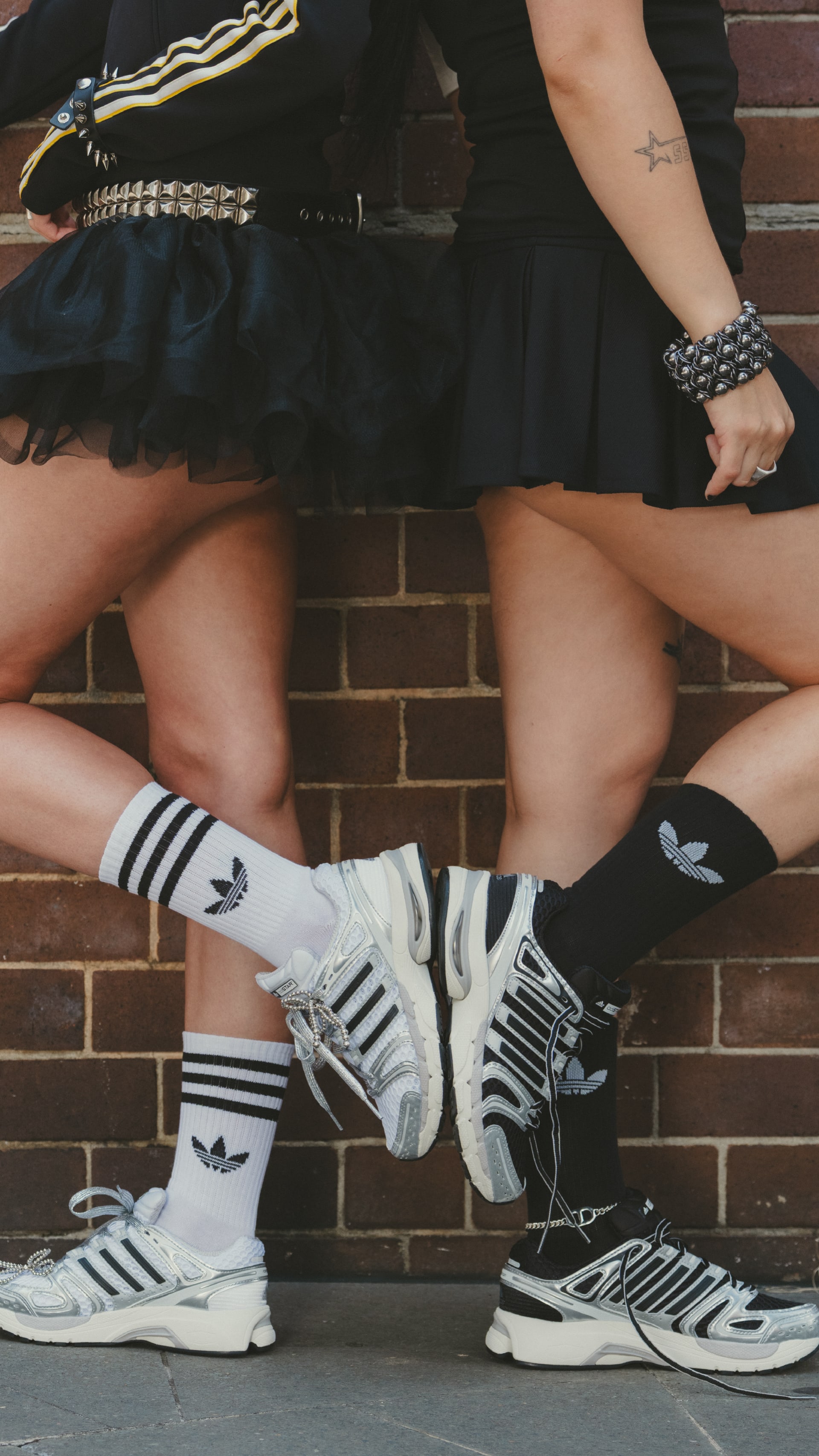 The image shows a person wearing black clothing, including a skirt, and Adidas sneakers, standing in front of a brick wall.