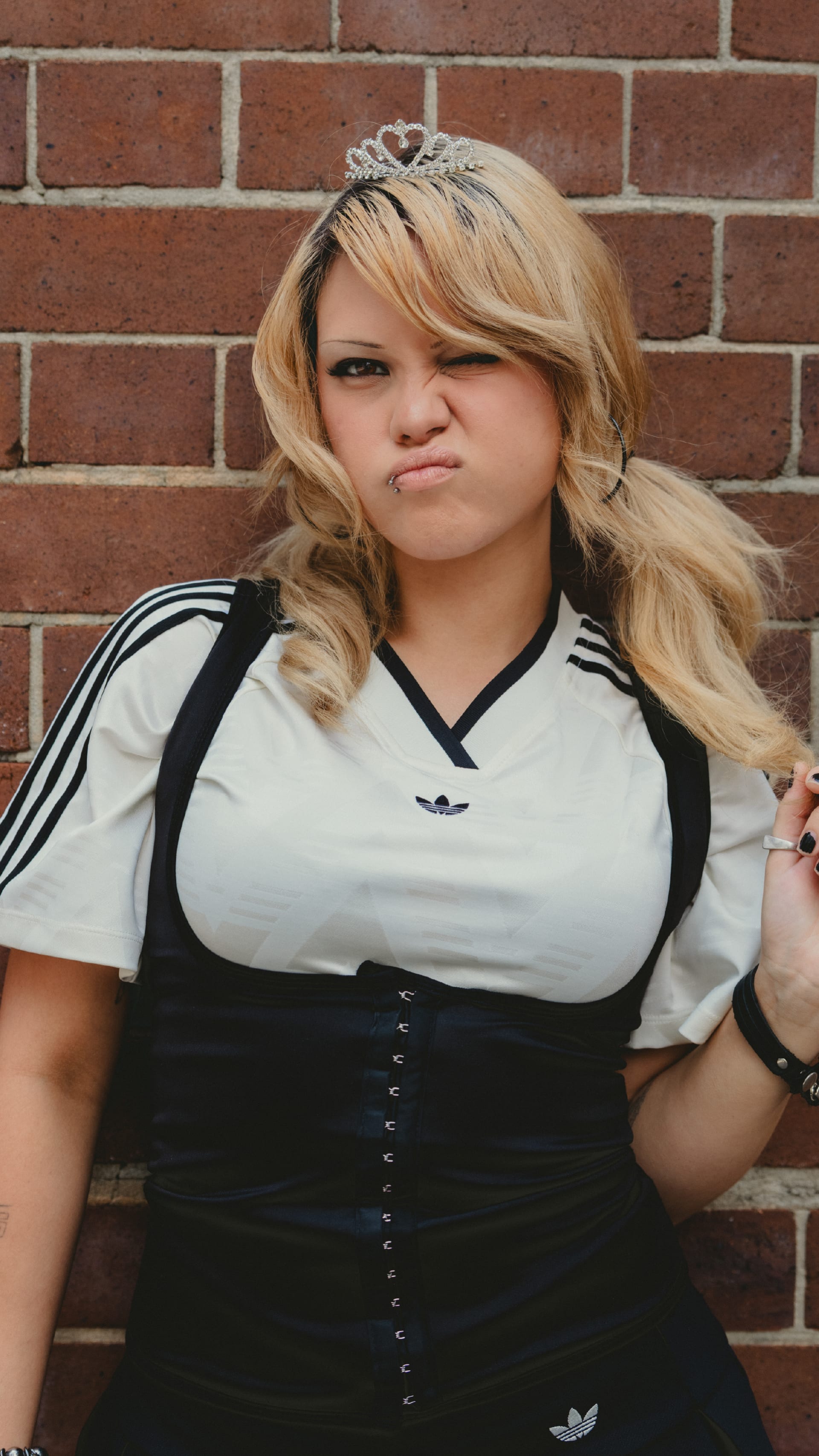 A young woman with blonde, curly hair stands in front of a brick wall, wearing a white and black athletic top.