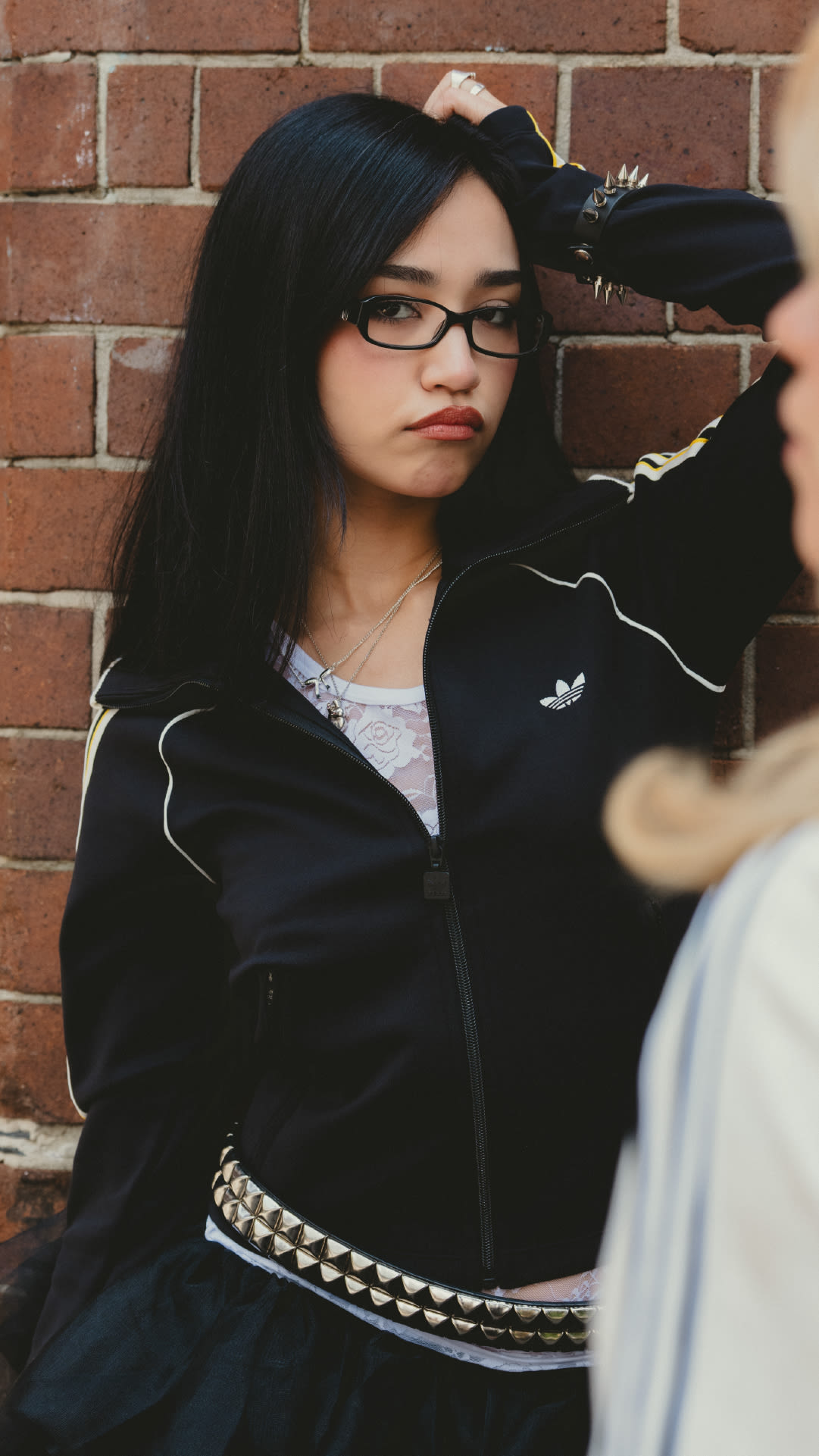 A young woman with dark hair and glasses stands in front of a brick wall, wearing a black jacket and a patterned skirt.