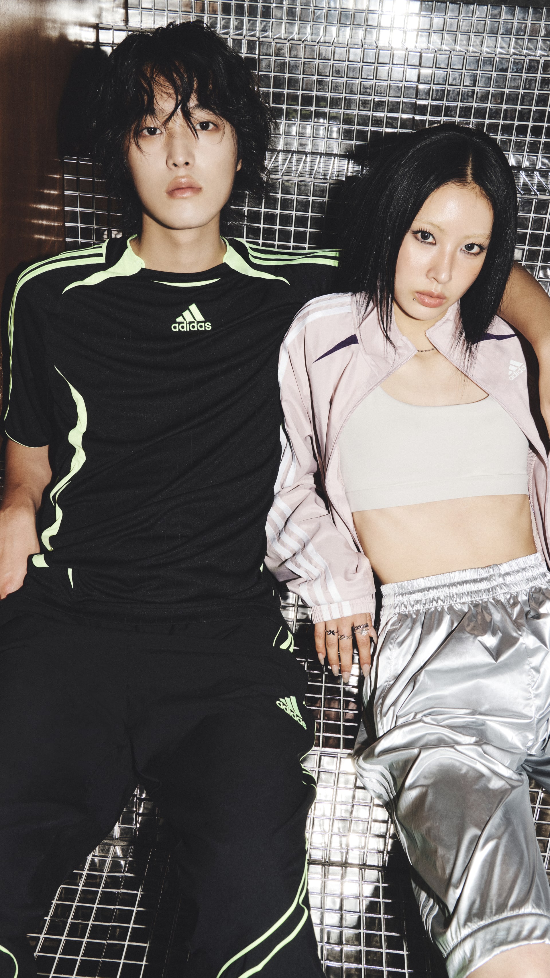 Two individuals, a man and a woman, are posing together in front of a metallic mesh backdrop. The man is wearing a black athletic shirt, while the woman is wearing a light-colored crop top and silver pants.