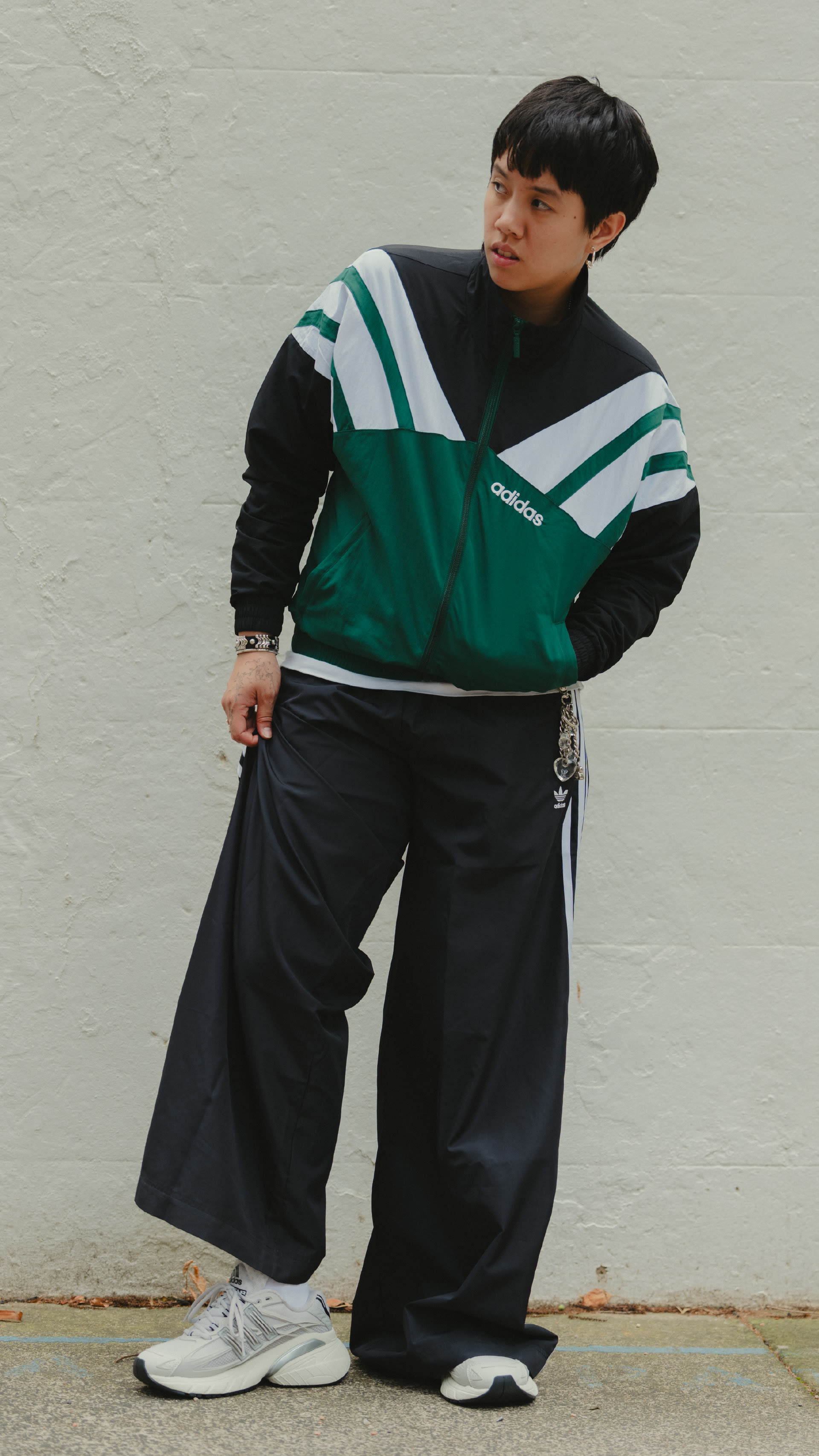 A young person wearing a green and black athletic jacket and pants, standing in front of a plain wall.