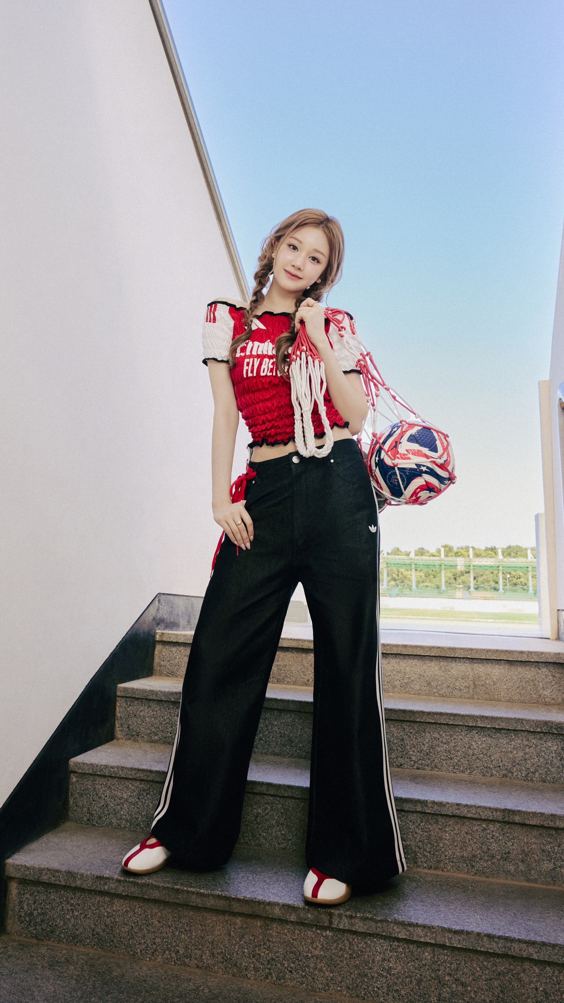 A young woman with long blonde hair stands on a set of stairs, wearing a red and white patterned top and black wide-leg pants, against a backdrop of a clear blue sky.
