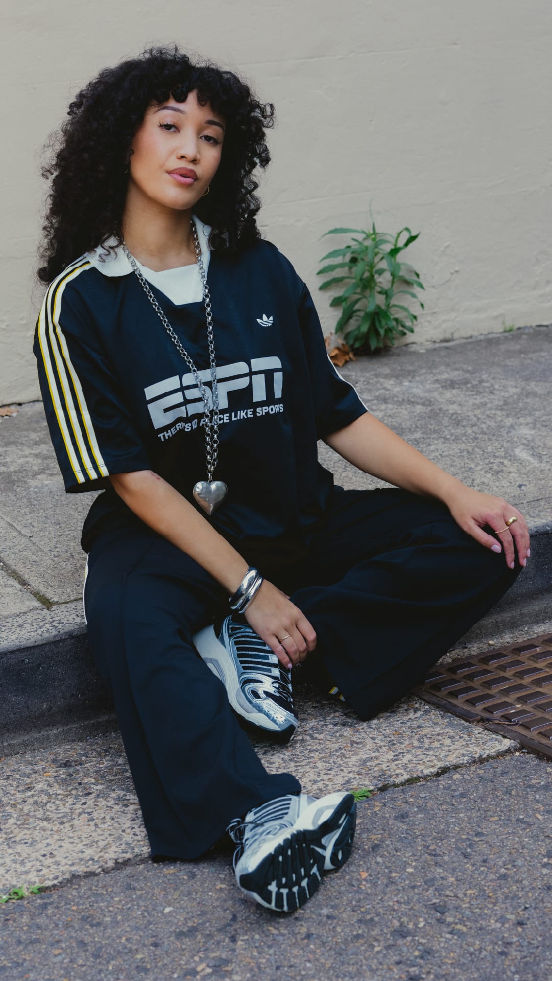 A young woman with curly dark hair sits on a concrete surface, wearing a black ESPN t-shirt and sneakers, with a plant visible in the background.