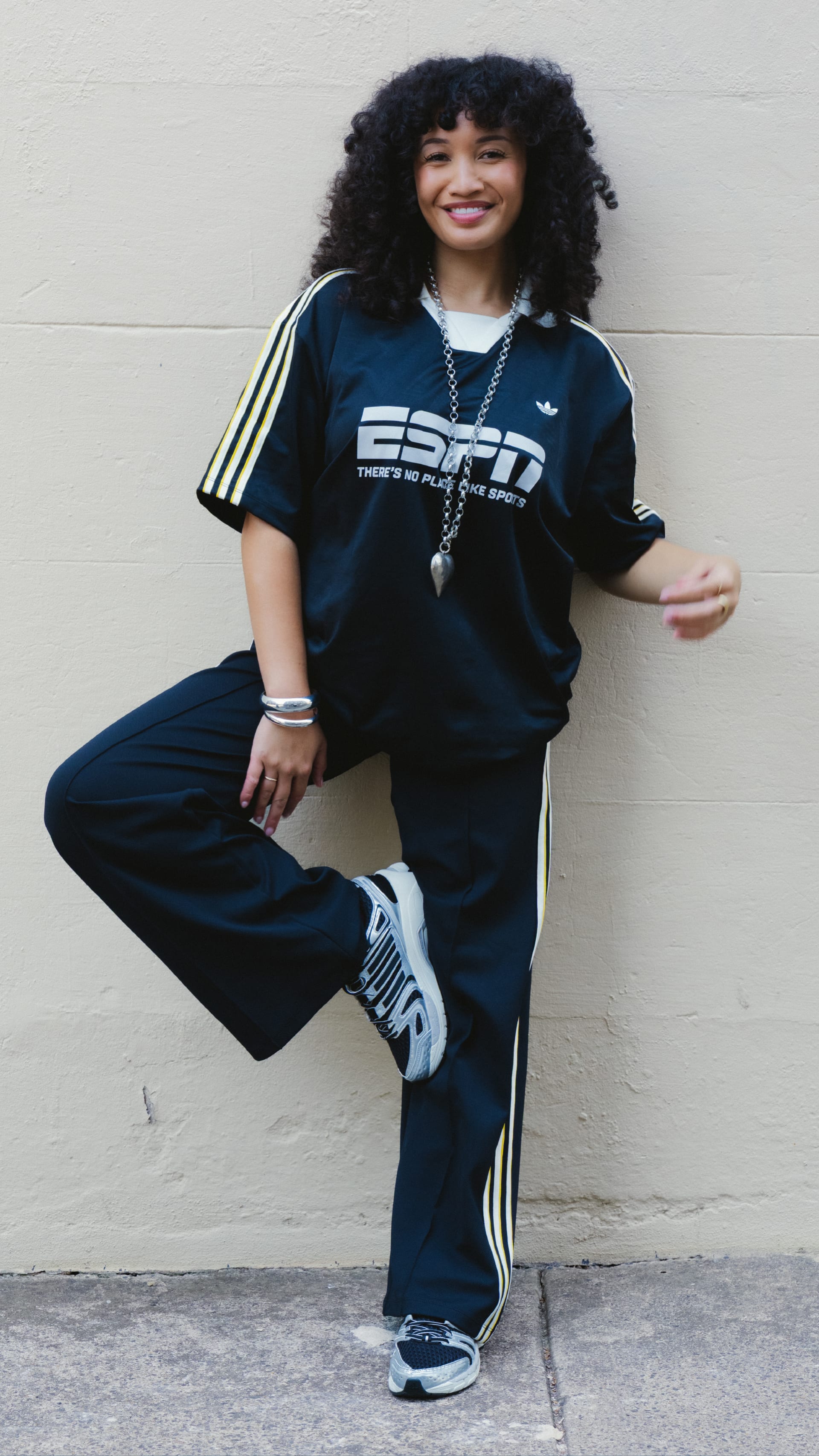 A young woman with curly hair wearing a navy blue Adidas tracksuit is standing against a white wall, smiling at the camera.