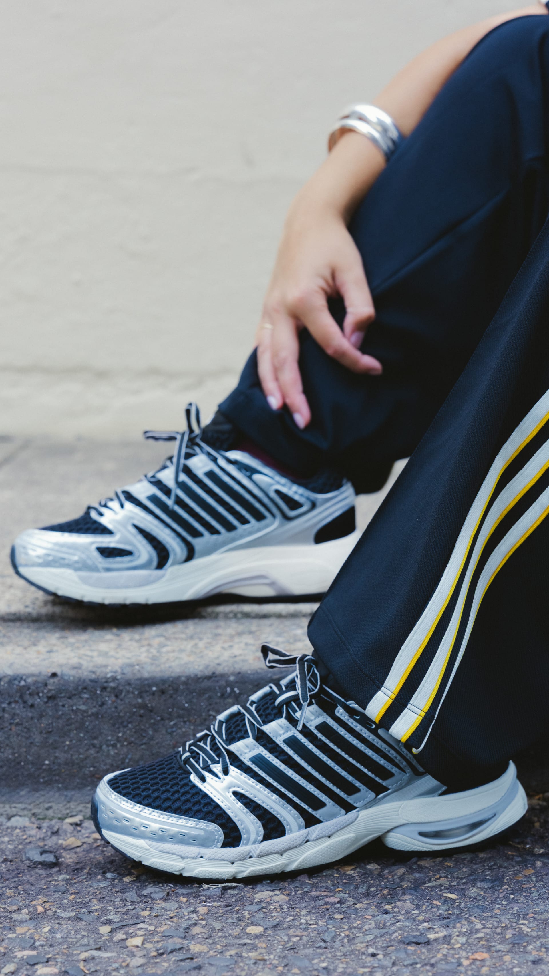 The image shows a person's lower body wearing navy blue pants and athletic shoes, sitting on a concrete surface with a blurred background.