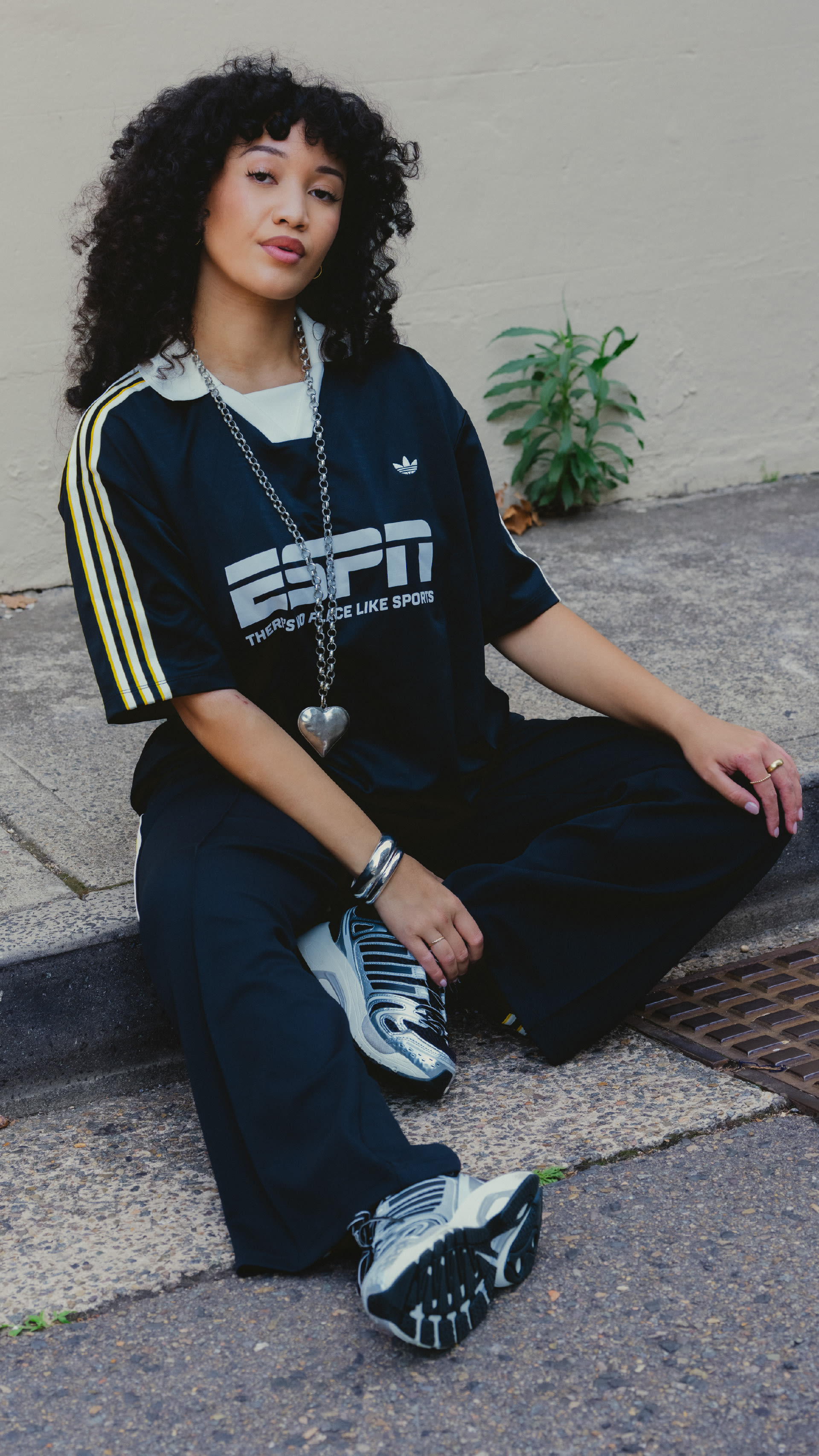 A young woman with curly dark hair sits on a concrete step, wearing a black and yellow sports jersey and sneakers, with a potted plant visible in the background.