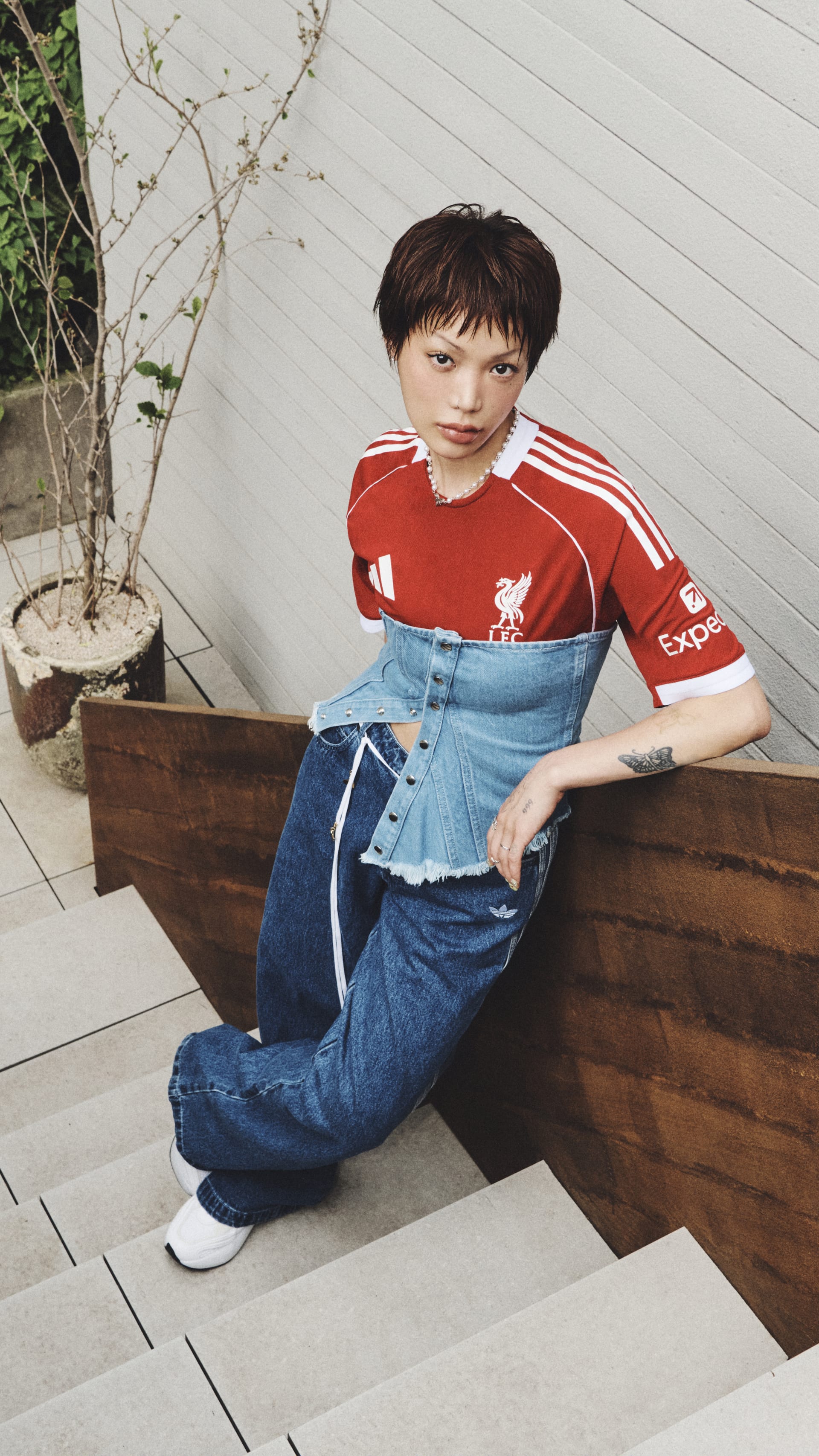 A young boy wearing a red sports jersey and jeans is sitting on a wooden structure in an outdoor setting with plants and greenery in the background.