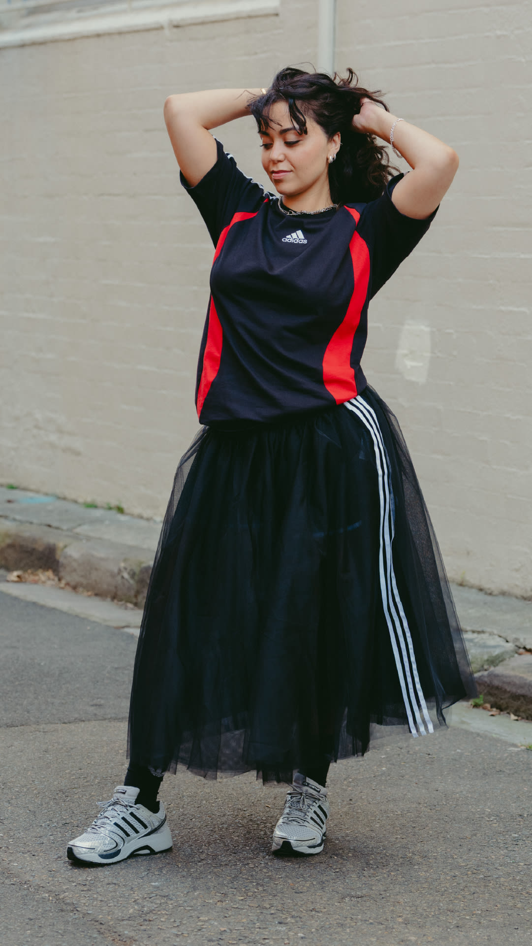 A young woman wearing a black and red athletic top, a black tulle skirt, and sneakers stands in an urban setting with a plain wall in the background.