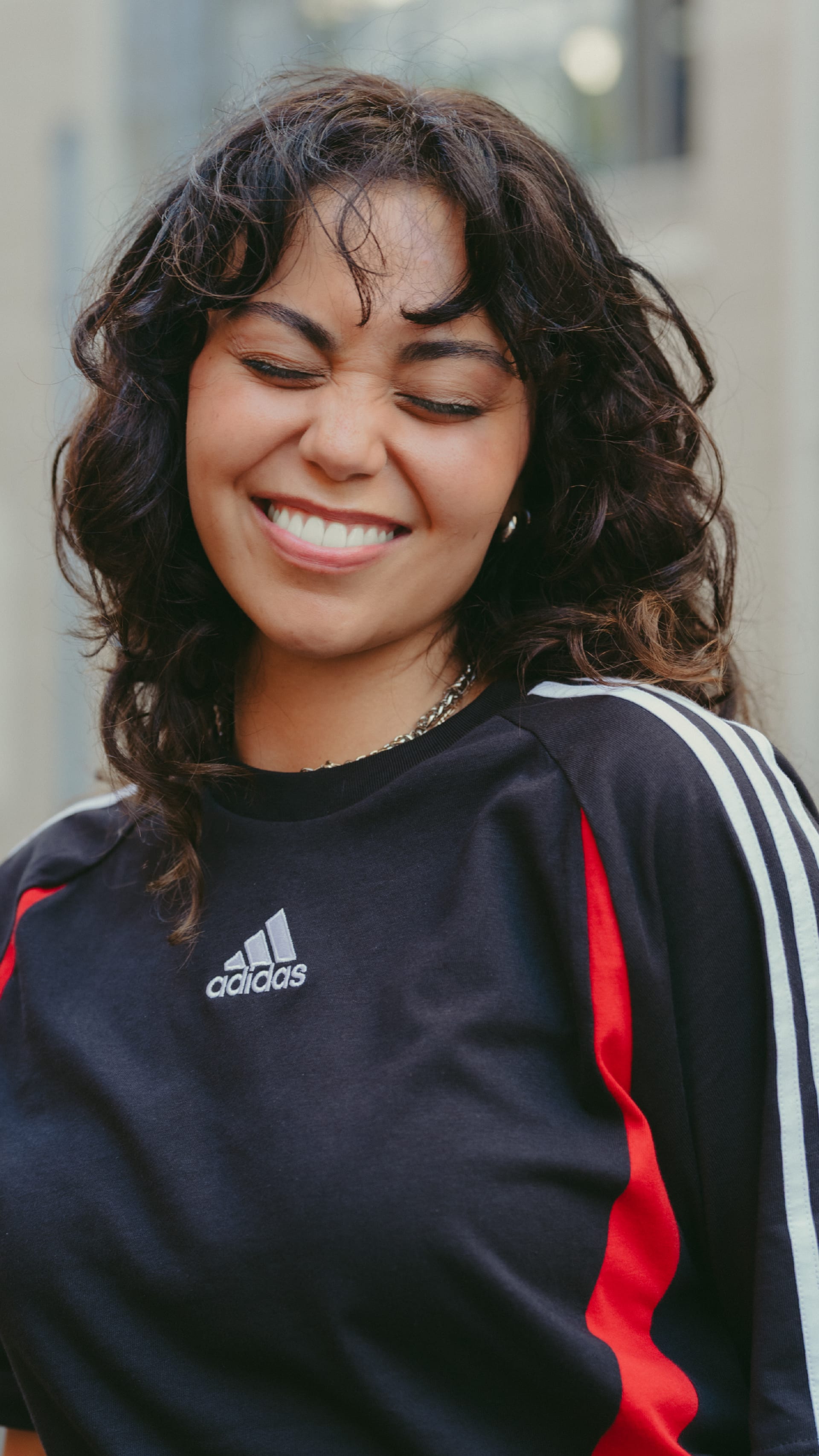 A smiling young woman with curly dark hair wearing a black and red Adidas athletic top stands in front of a blurred background.