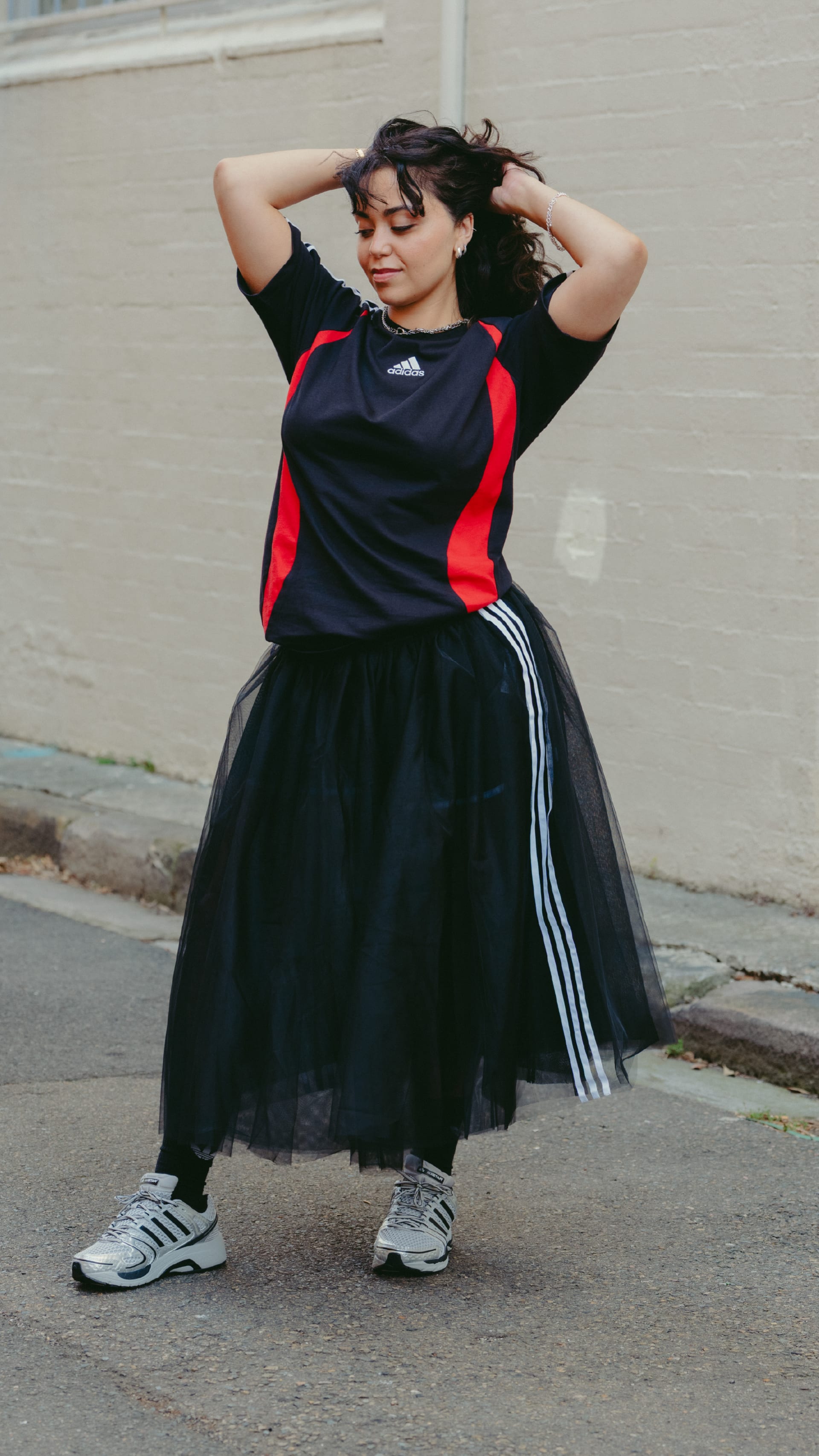 A young woman wearing a black and red athletic top and a black tulle skirt stands in front of a plain wall, her hands raised to her head as she appears to be adjusting her hair.