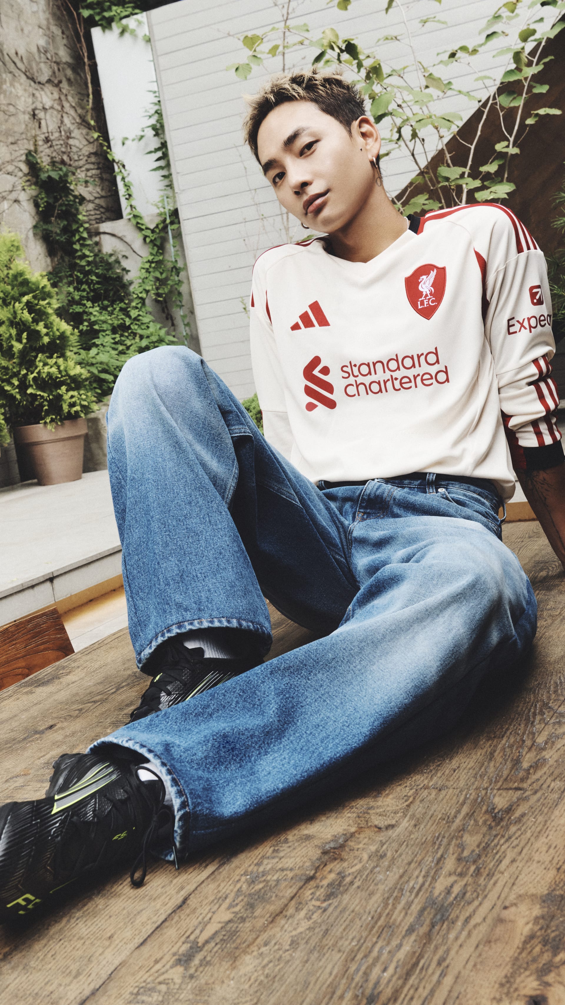 A young man wearing a Liverpool football club jersey is sitting on a wooden floor, surrounded by potted plants and a white wall in the background.