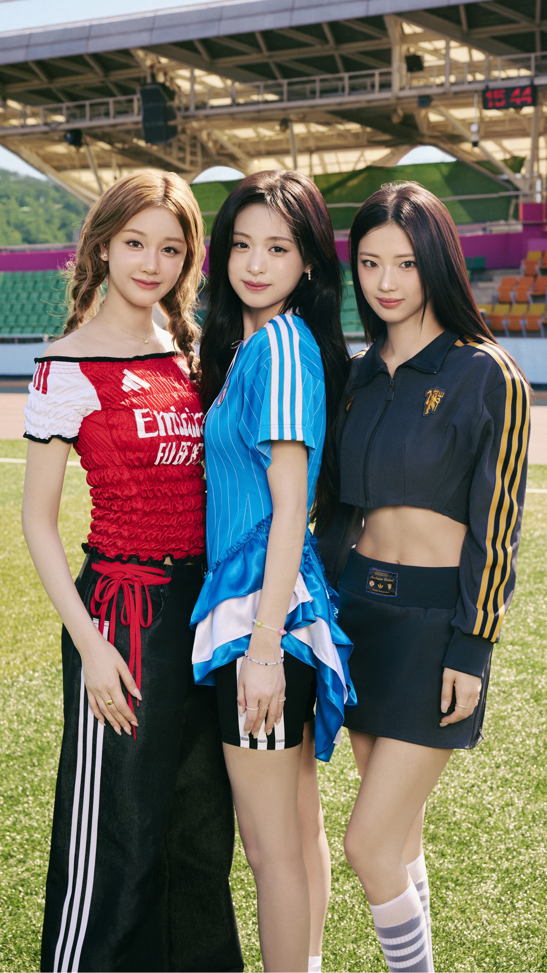 Three young women in colorful athletic attire stand together on a sports field with stadium seating visible in the background.