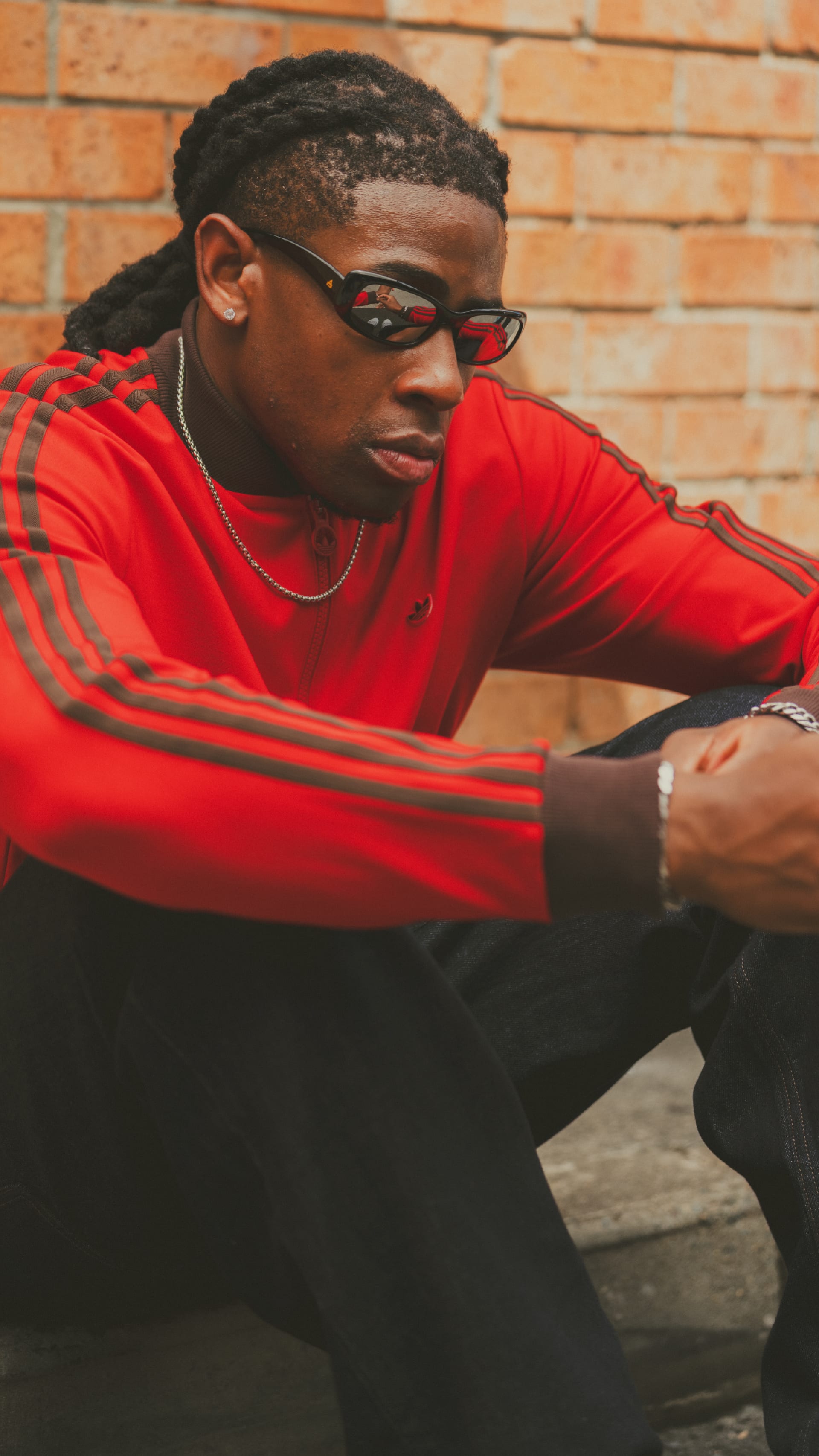 A young man with braided hair wearing a red shirt and glasses, sitting against a brick wall background.