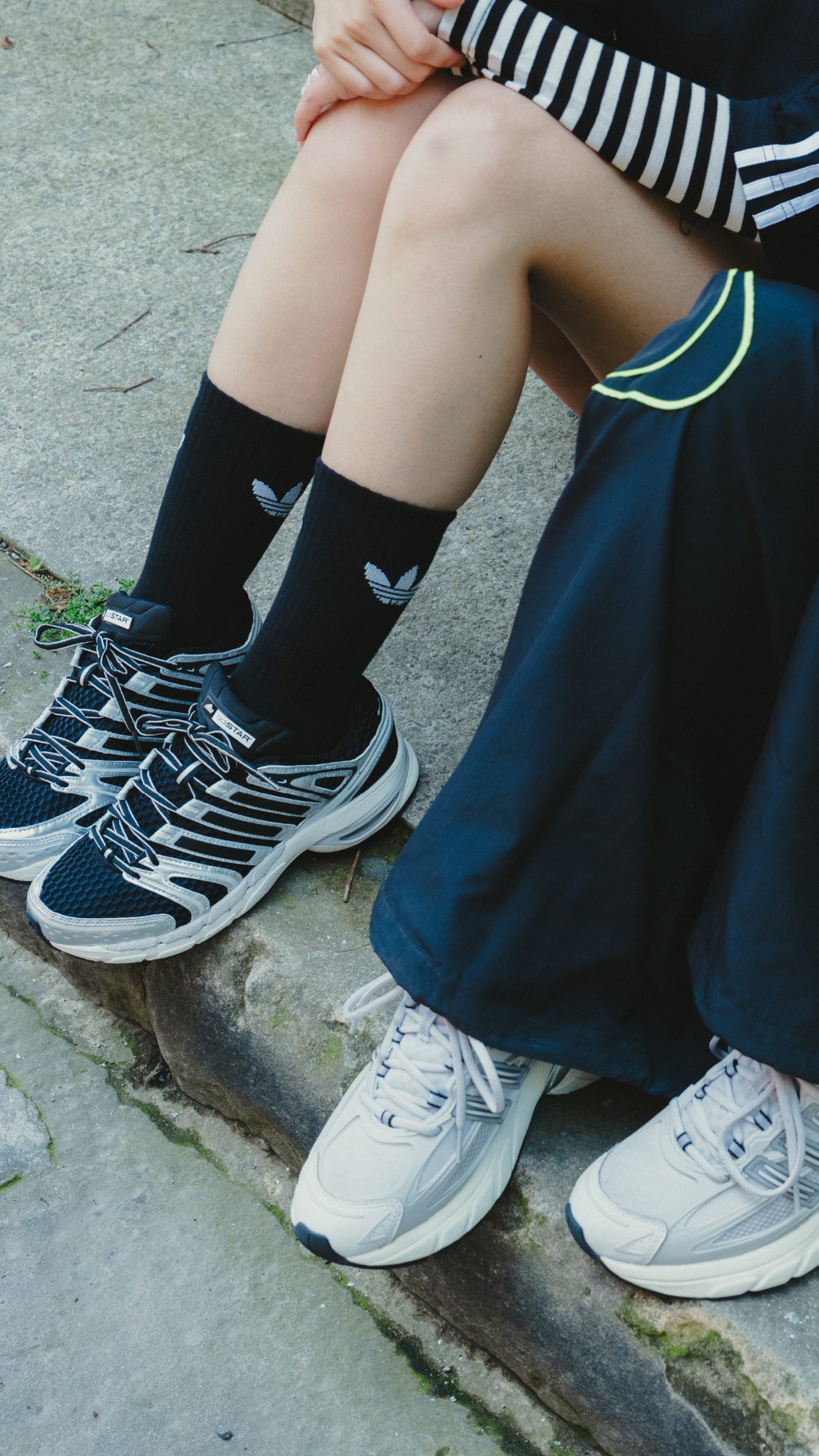 The image shows a person's legs and feet, wearing striped socks, black sneakers, and a blue skirt or dress, sitting on a concrete surface.