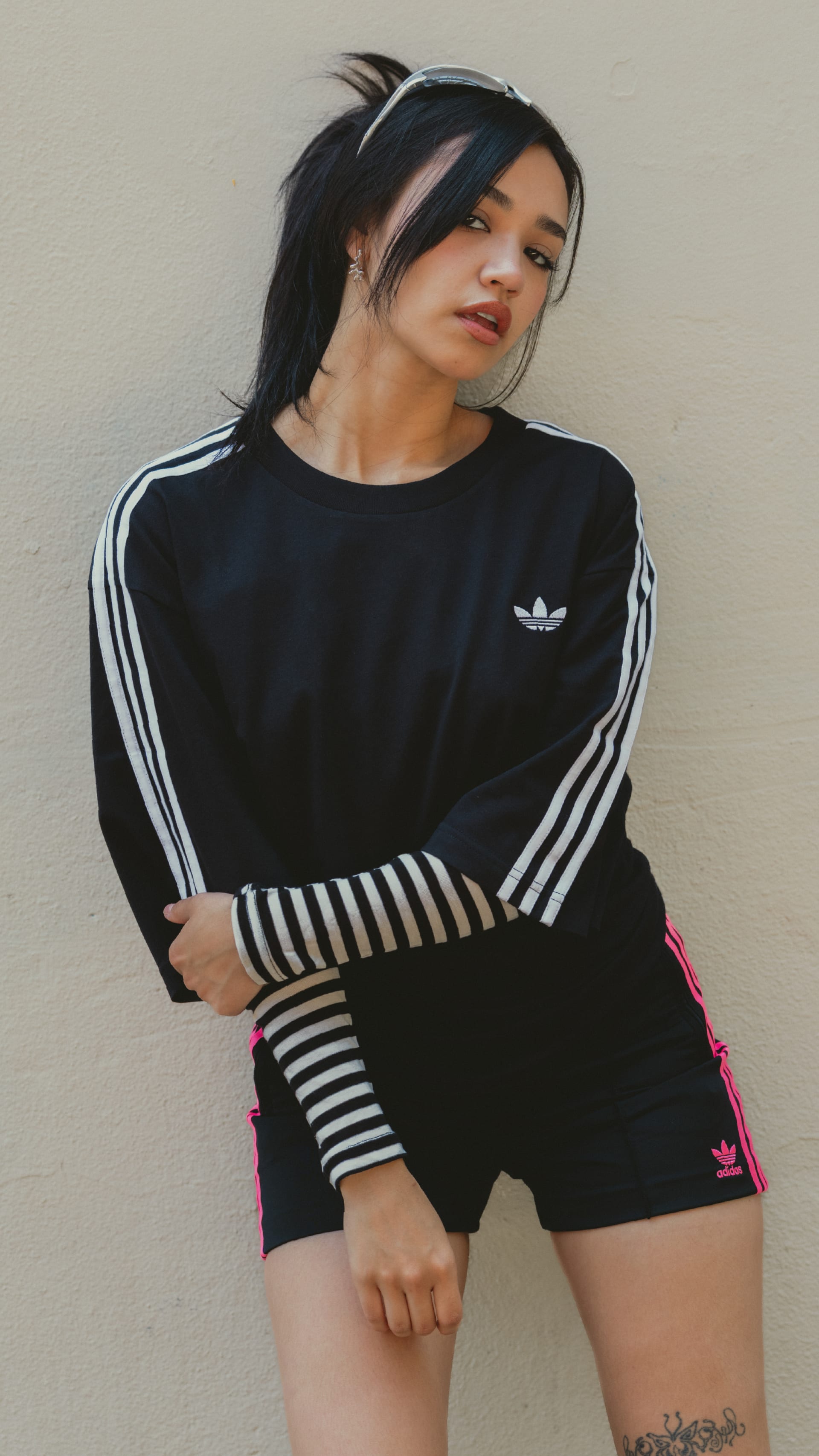 A young woman wearing a black Adidas top with white stripes stands against a plain background, her arms crossed and a thoughtful expression on her face.