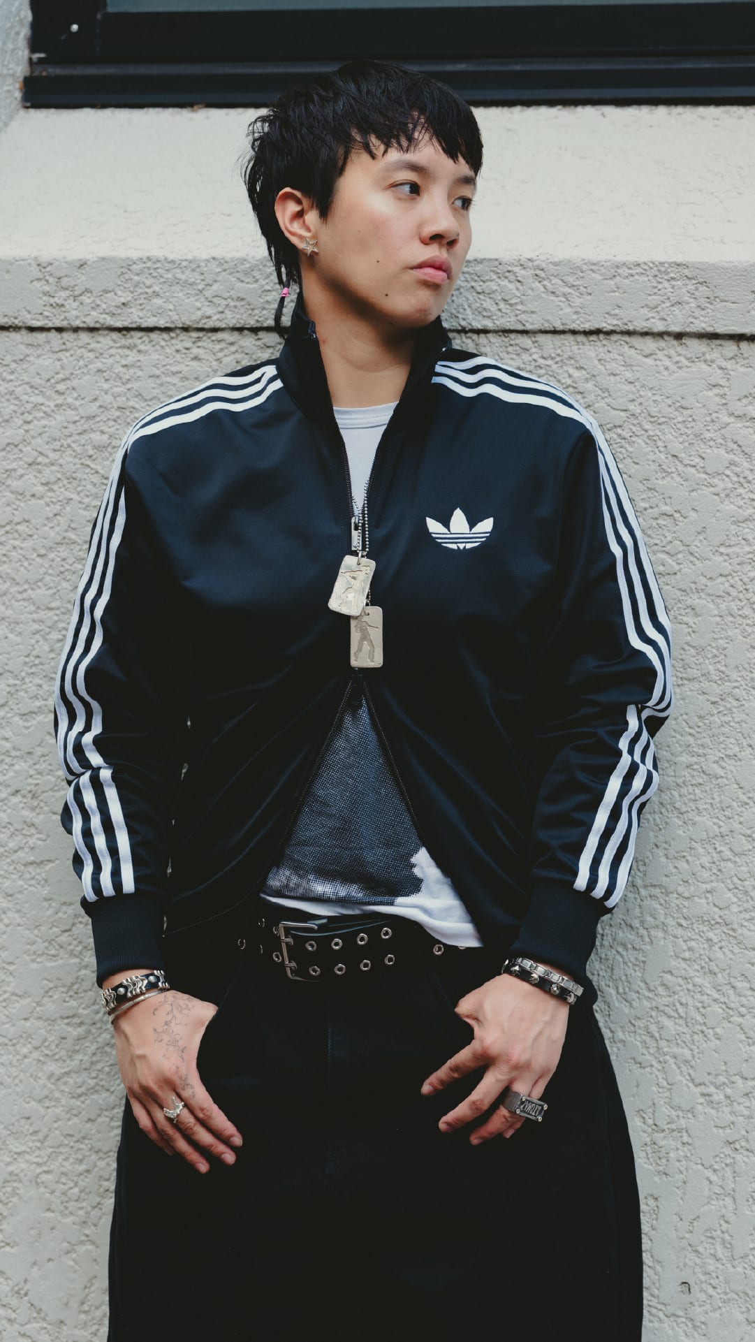 A young person wearing a black Adidas jacket with white stripes stands in front of a plain white wall.