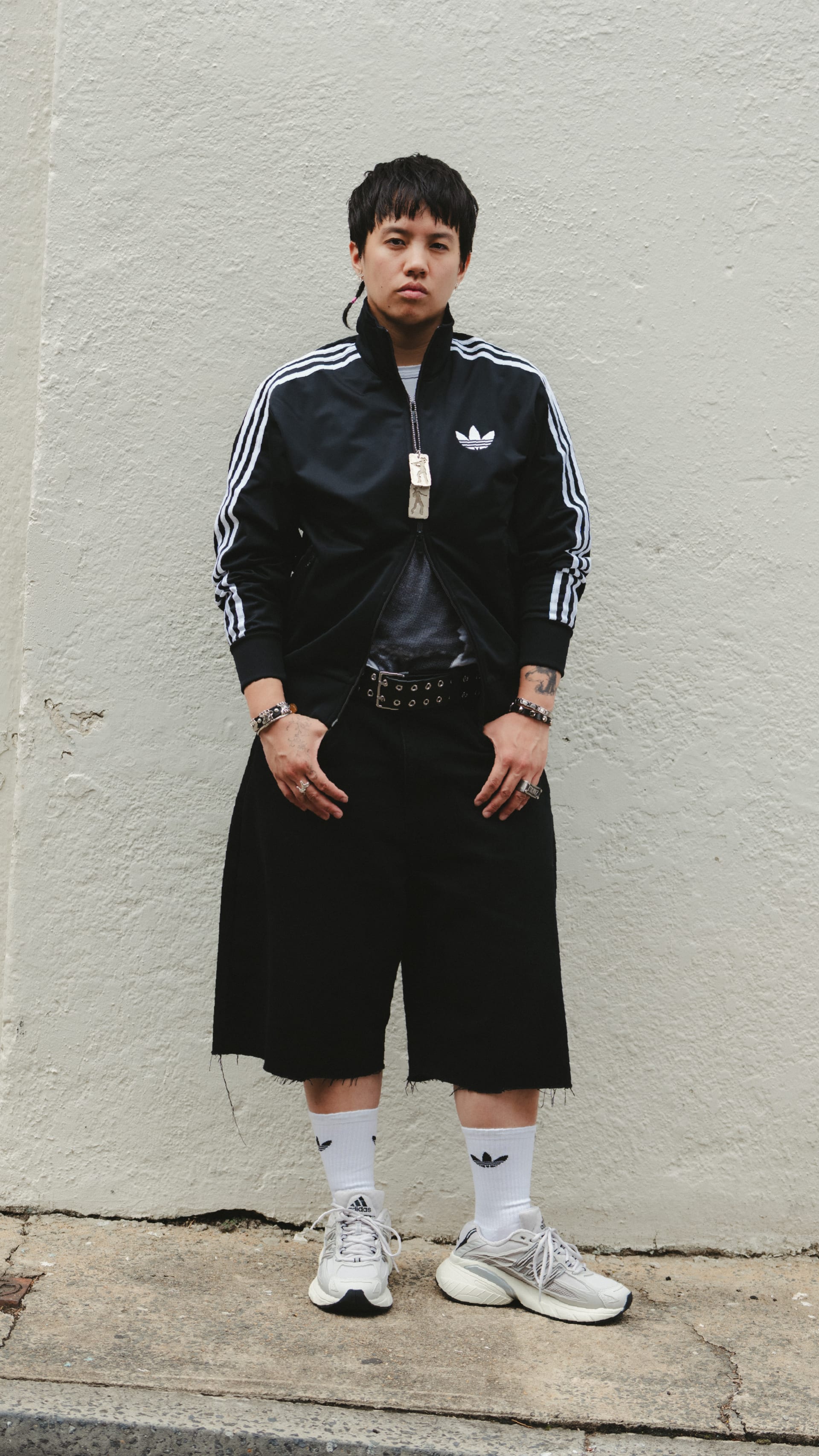A young person wearing a black tracksuit and sneakers stands in front of a plain white wall.