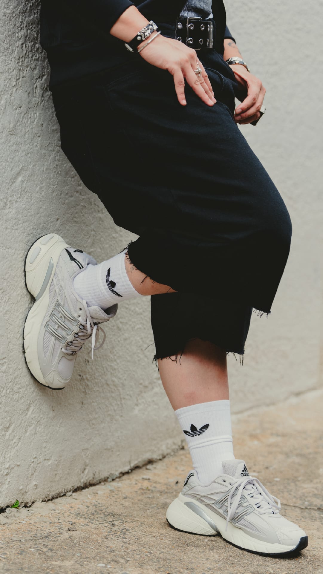 The image shows a person wearing black clothing, including a skirt or dress, and white sneakers, sitting on the ground against a plain wall.